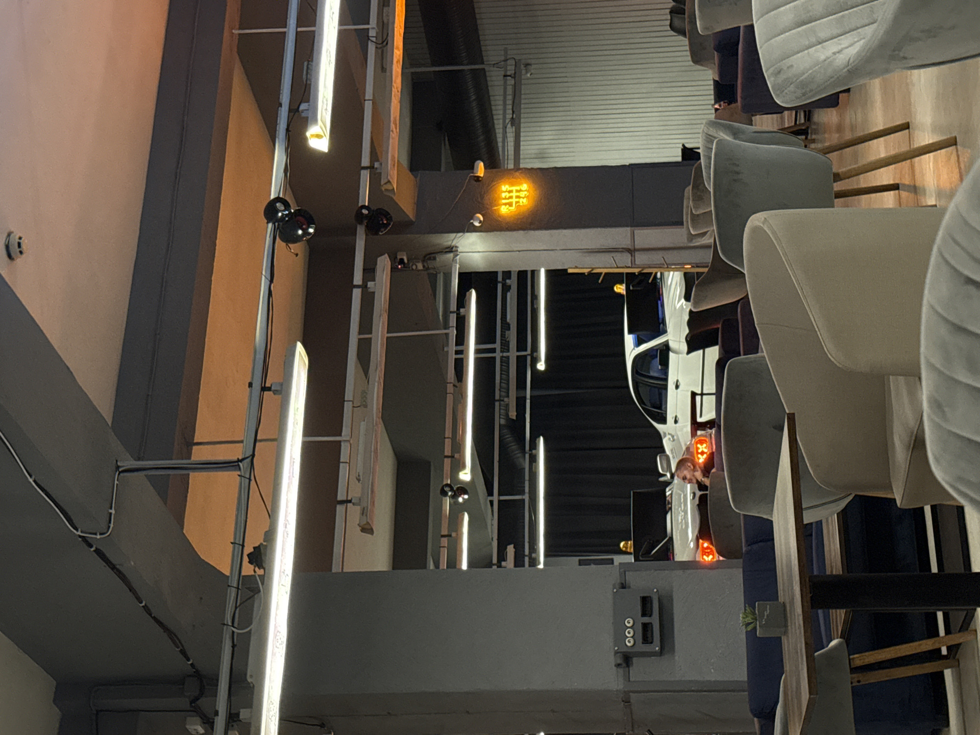 This is a medium shot, eye-level photograph of a dimly lit indoor space that appears to be a lounge or cafe. The scene is dominated by concrete pillars and ceilings, with exposed electrical conduits and fluorescent lighting fixtures. In the background, a white car with its brake lights illuminated is partially visible, with a person in the driver's seat. The car is positioned in front of dark drapes. To the right of the car, a neon sign displaying a gear shift pattern is illuminated in orange. The foreground is filled with various upholstered chairs and a table with a potted plant. The furniture varies in color and style, with some chairs in shades of navy blue, beige, and light gray. The overall ambiance is moody and modern. The location is identified as Unknown, Unknown.