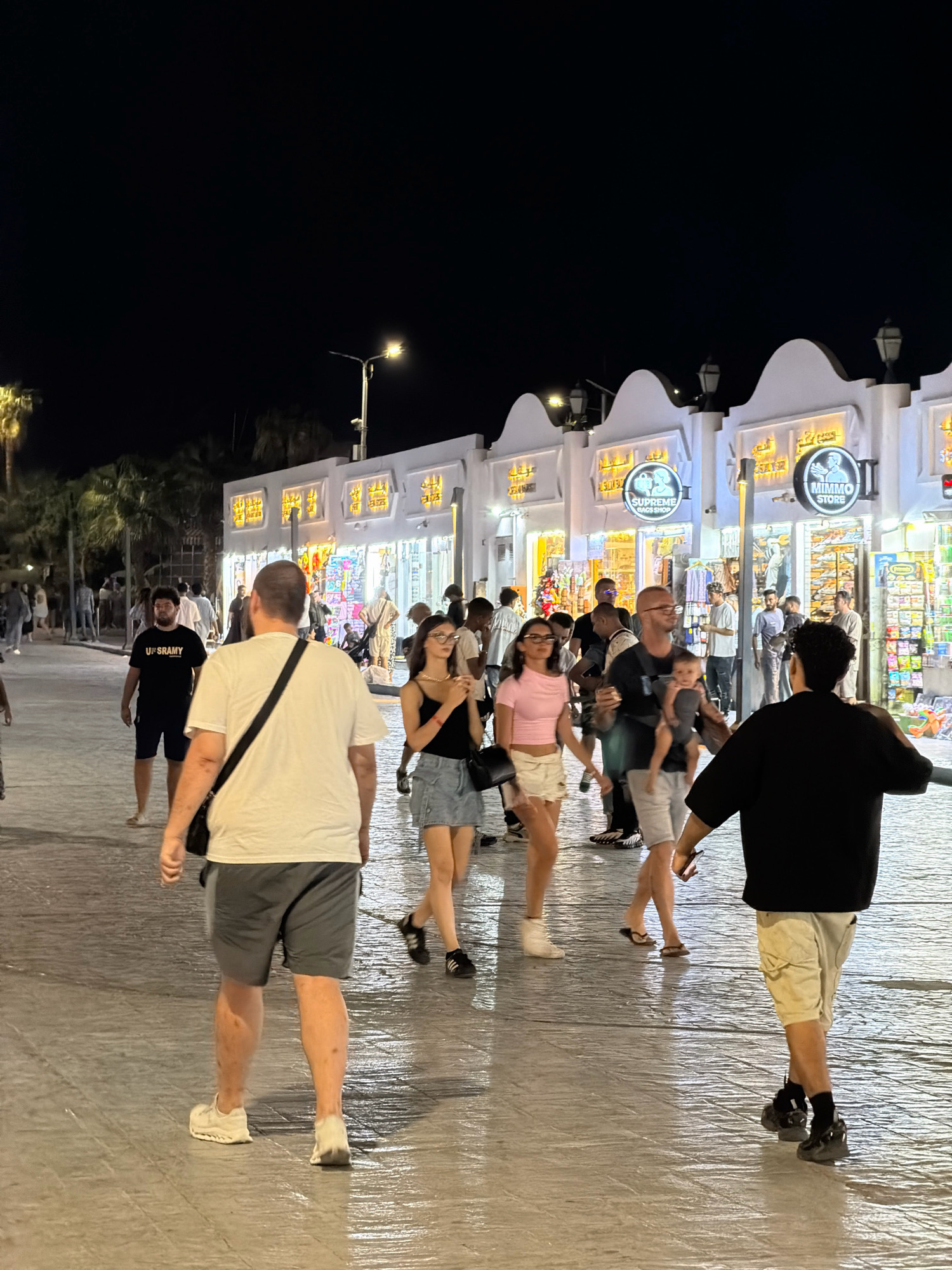 A nighttime street scene in Sharm El Sheikh, Egypt, shows a paved walkway lined with shops. Several people are walking along the walkway. In the foreground, a man wearing a white t-shirt and gray shorts walks away from the camera. To his left, a man in a black t-shirt with "UP SRAMY" written on it walks towards the camera. To the right of the man in the white t-shirt, a woman in a pink top and denim skirt walks towards the camera, and a man carrying a baby in a sling walks past her. Further back, more people are visible, and the illuminated storefronts display various goods. The street is wet, reflecting the lights. Palm trees are visible in the background.