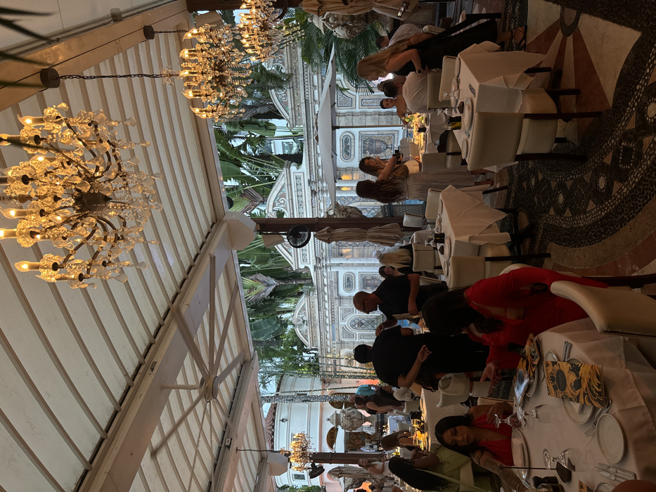 The image depicts an upscale outdoor dining area at The Villa Casa Casuarina, formerly the Versace Mansion, located on Ocean Drive in Miami Beach, United States.
The scene is set under a large, retractable or permanent light-colored awning ceiling, from which multiple ornate crystal chandeliers hang, casting a warm, golden light. A large, industrial-style ceiling fan is also visible. The ground is covered with a patterned mosaic tile floor.
In the background, the distinctive classical architecture of the Versace Mansion is prominent, featuring white stucco walls adorned with intricate relief work, pilasters, and friezes, consistent with the "Thousand Mosaic Pool" area. Lush tropical foliage, including tall palm-like trees, rises above the mansion walls. A large decorative urn is visible near a circular tower structure to the left. A wall-mounted fan is affixed to a column.
The dining area is furnished with numerous square and rectangular tables, draped with white tablecloths. Each table is set with white plates, glasses, and cutlery. Several tables feature distinctive menus with gold and black patterns. Upholstered dining chairs, primarily light in color, are arranged around the tables.
Approximately twenty to thirty individuals are present, engaging in various social activities. In the foreground, two women are seated at a table. One, on the far left, wears a red top and looks to her left, with a prominent tattoo on her left arm. Another woman in a red top/dress is partially obscured, reaching or gesturing towards the table. In the mid-ground, a man in a black shirt stands with his back to the viewer, conversing with another man in a black polo shirt and eyeglasses. Towards the right, groups of people stand and sit around tables; a woman in a black dress walks away from the camera, and another in a striped dress stands near a table. A person is partially visible in the water of a pool in the mid-right background, behind some diners. The overall attire suggests evening wear ranging from casual to semi-formal. The lighting indicates dusk or evening hours, with artificial illumination creating the primary light source.