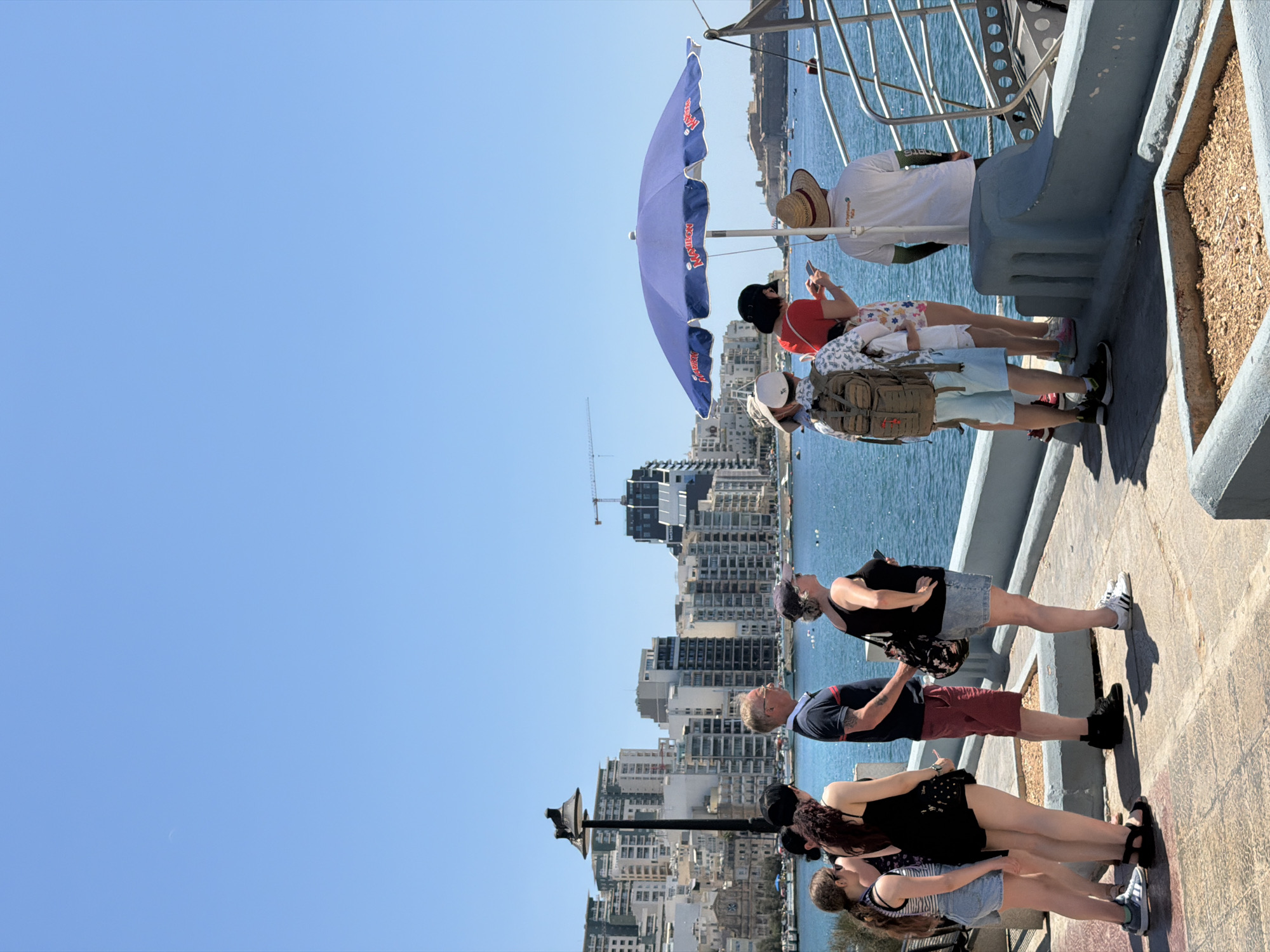 This media file captures a group of people standing on a promenade with a view of a city skyline and the sea. The scene is set on a bright, sunny day, suggesting it is midday. The architecture of the buildings in the background indicates a Mediterranean location, with Sliema, Malta, identified as the context.
In the foreground, several individuals are casually dressed, some wearing hats and sunglasses. One person is holding a mobile phone, and another is looking up at the sky. A large, blue umbrella is open, providing shade for some of the people. A backpack is visible, suggesting travel or a day out. The people appear relaxed and are observing the surroundings.
In the middle ground, the cityscape is characterized by numerous multi-story buildings clustered together along the waterfront. The sea is a prominent feature, calm and blue, with a few boats visible in the distance. The promenade itself is paved with stone.
Notable details include the clear blue sky, indicating good weather, and the sunshine casting shadows. The overall mood seems peaceful and leisurely, with individuals enjoying the scenery and each other's company. The visible text on the umbrella reads "RED BULL," a well-known brand.