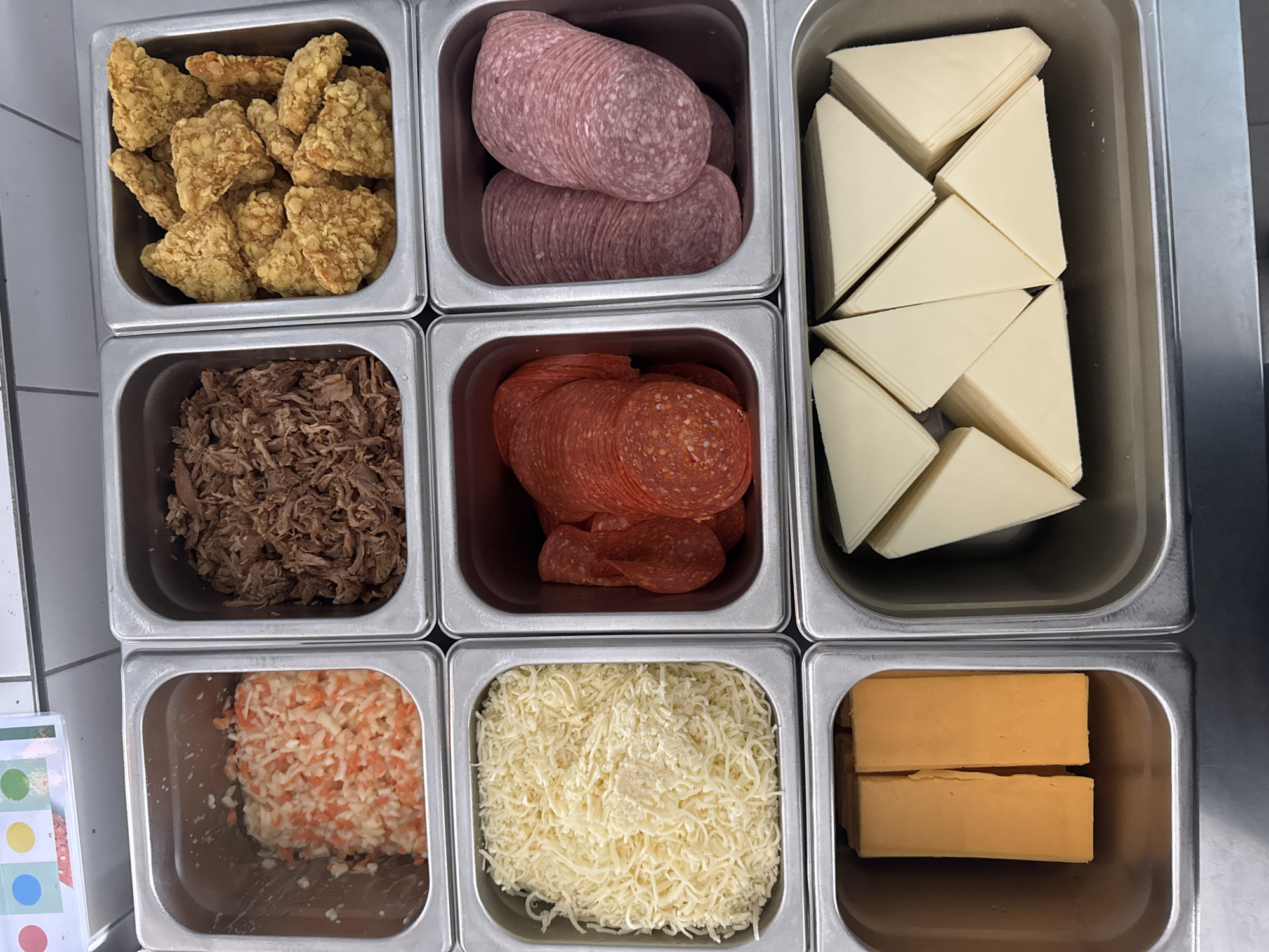 An overhead shot shows a food preparation station with nine stainless steel containers filled with ingredients. In the top row, the left container holds a mixture of shredded cabbage and carrots. The middle container contains shredded pulled pork. The right container is filled with crispy, golden-brown fried chicken pieces.
The middle row features three containers. The left container is full of finely shredded white cheese. The center container displays a stack of thinly sliced red pepperoni. To the right, another container holds two rolls of sliced cured sausage.
The bottom row contains two containers. The left container has two rectangular blocks of orange cheddar cheese. The right container is filled with sliced pale yellow cheese, cut into triangular wedges and stacked. The entire setup appears to be organized for food assembly, likely for dishes like pizzas or sandwiches. The background shows a white tiled wall and a small sign with colored circles.