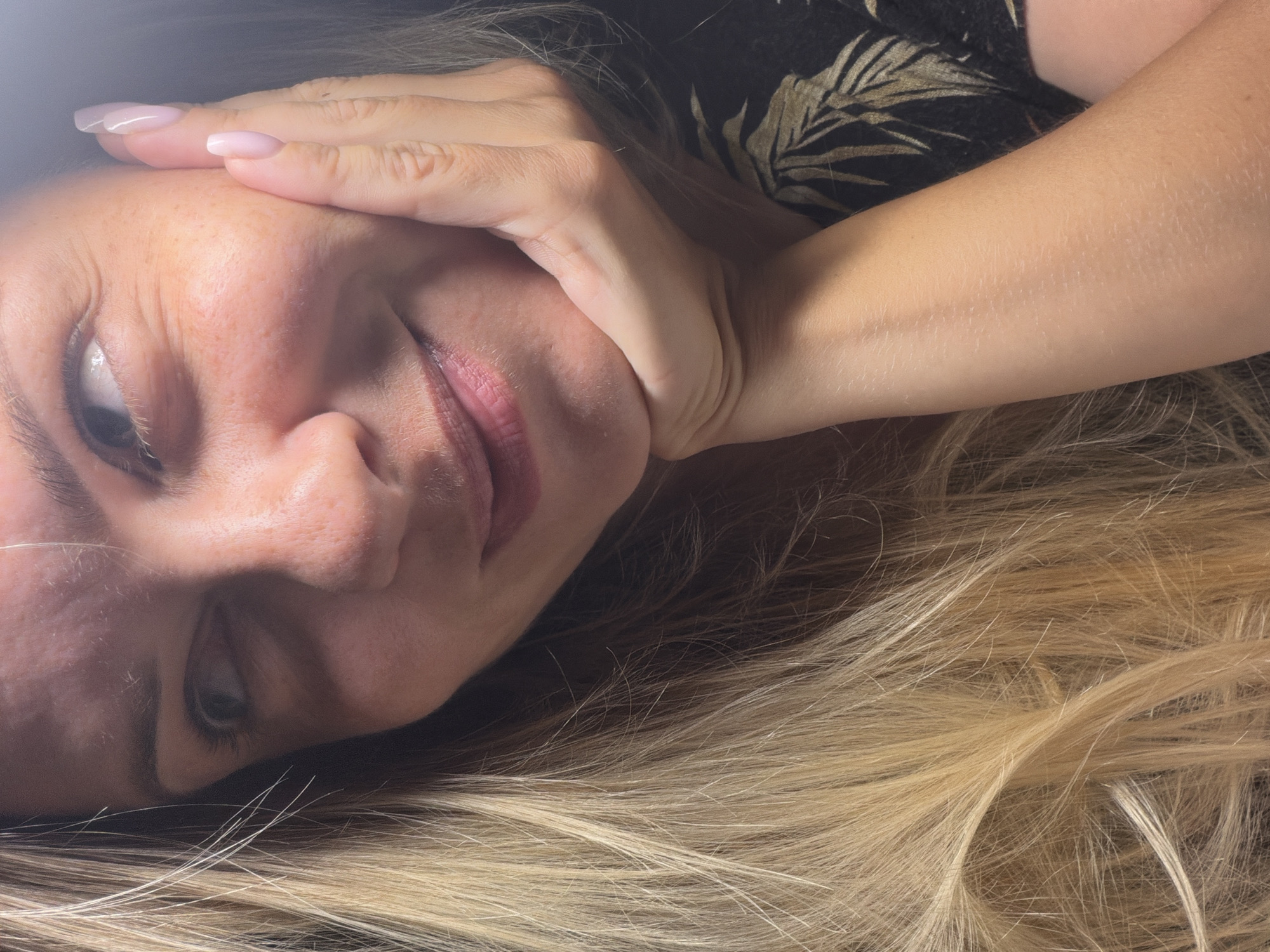 A close-up, low-angle selfie shows a blonde woman with light skin and freckles. Her right hand is propped on her chin, with her fingers touching her cheek. She has light pink, oval-shaped fingernails. She is looking to her left with a slight smile, and her mouth is slightly open. Her blonde hair is visible on the left side of the frame. She is wearing a dark shirt with a pattern of gold leaves. The background is dark. The image does not contain any information about the location in Votkinsk, Russia.
