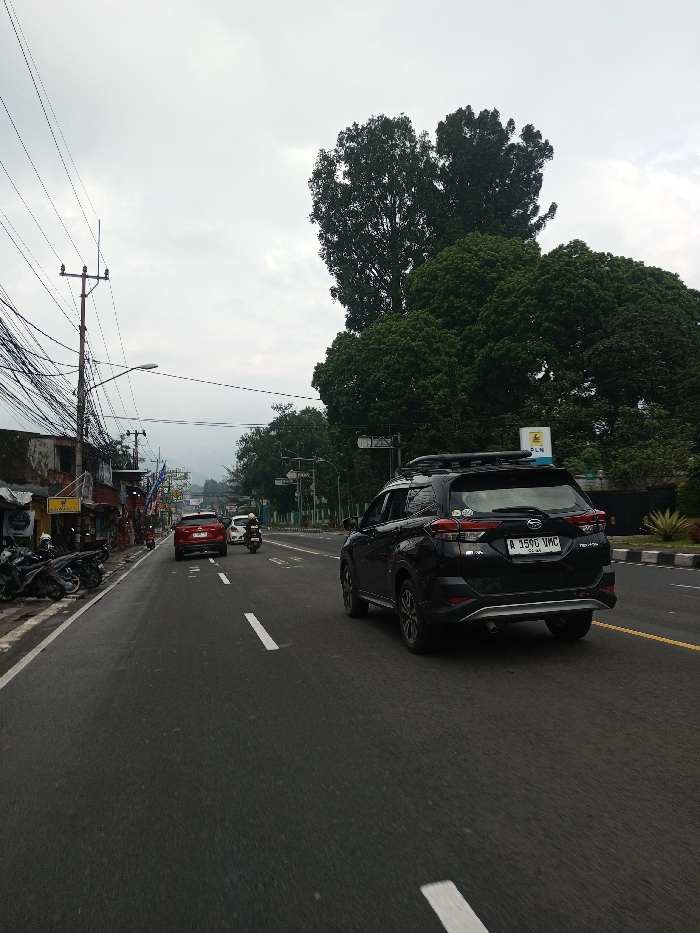 This image depicts a street scene captured on an overcast day in Dawathang_Dorjibi_ Kashingtsawa, Bhutan. The perspective is from a low angle on the asphalt road, looking forward down a multi-lane thoroughfare.
The road features white dashed lane markings, indicating at least two lanes of traffic moving away from the viewer. A black Daihatsu Terios/Toyota Rush SUV with a roof rack is in the rightmost lane, displaying the license plate "A 3590 UMC" on its rear. Ahead of it, a red sedan proceeds in the same lane. To the left of the red sedan, a single individual rides a white scooter or motorcycle, also moving away from the camera. Several motorcycles are parked at an angle on the left side of the road, adjacent to a concrete curb.
The left side of the scene is lined with low-rise buildings exhibiting worn facades, some with corrugated metal awnings. A prominent yellow rectangular sign on one building reads "SERUASLI". Multiple utility poles rise from the left, heavily laden with a dense network of black overhead wires.
On the right side of the road, a concrete curb separates the roadway from a landscaped median strip featuring short green shrubs and a substantial treeline composed of tall, mature trees with dark green foliage. A yellow and blue rectangular sign, partially obscured by trees, displays the letters "PLN". In the distant background, hazy mountains or hills are visible beneath a uniformly grey, cloudy sky. No other discernible human figures are present apart from the scooter rider.