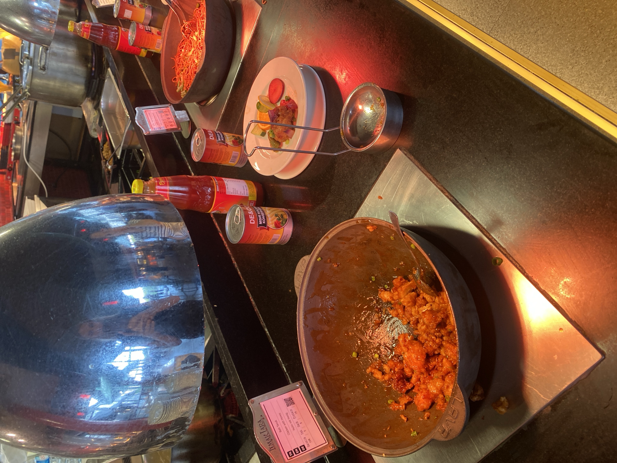 The image depicts a food buffet in a restaurant, possibly at an all-inclusive hotel, in Aksu, Turkey. The buffet line features various food items, including what appears to be fried chicken in a large pot, a plate with a small portion of food and fruit garnish, and a bowl of noodles. Several bottles of sauces and cans of food are also visible on the counter. In the background, kitchen equipment such as pots and a stove are present. The lighting suggests an indoor setting, possibly a dining area. There is a reflection in a large metallic dome, showing a woman taking a photo of herself with a phone. The overall scene is one of a self-service dining experience.