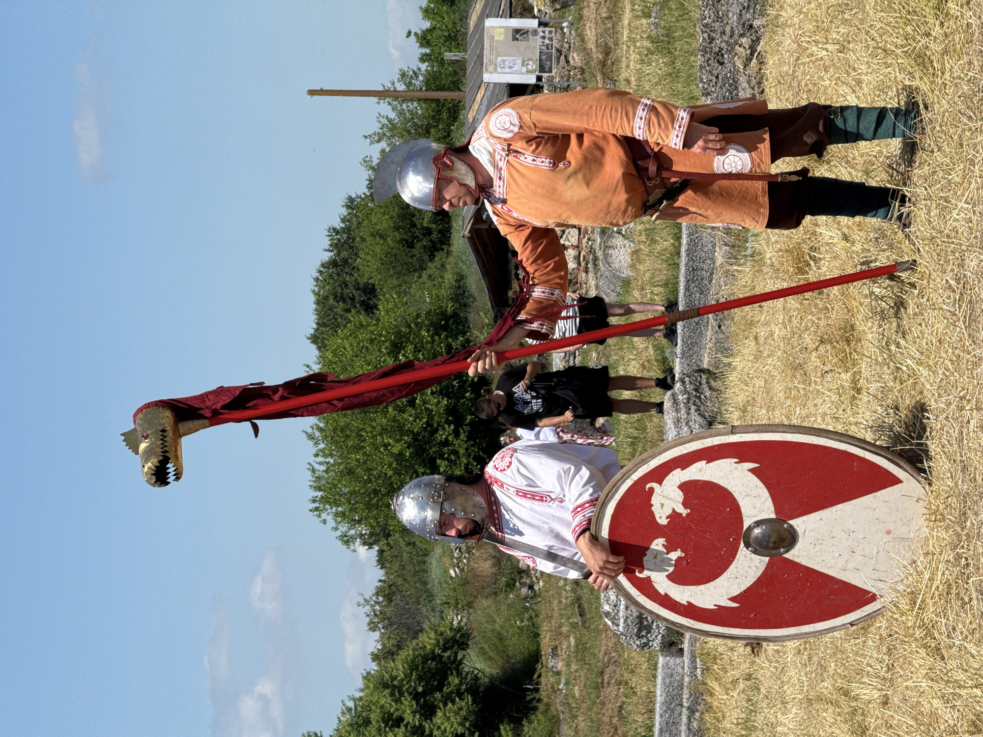 The image depicts an outdoor scene at an archaeological site in the city of Gigen, Bulgaria, under a bright blue sky with scattered white clouds. Two individuals in historical military costumes are standing on dry, light-brown grass, surrounded by remnants of stone structures and green foliage.
The individual on the left is a Caucasian male wearing a polished silver-colored conical helmet, a white tunic with intricate red and white geometric embroidery on the collar, sleeves, and chest, and a brown belt. He holds a large, round shield, predominantly red with a central metallic boss. The shield's design features two opposing white stylized dragon or serpent heads with yellow eyes, forming a C-shape around the boss, with a white triangular base below it. A dark-colored object, consistent with a sheathed sword, is visible at his left hip. He stands with his body slightly angled to the right, looking forward.
The individual on the right, also a Caucasian male, wears a polished silver-colored helmet with a distinct ridge and crest, an orange-brown tunic with red and white embroidered patterns on the sleeves and shoulders, dark brown knee-length breeches, and green leg wraps secured by brown straps. A small knife or dagger is attached to his brown belt. He holds a tall, red pole-standard topped with a golden, stylized dragon head that has prominent teeth and red eye-like accents. A red fabric streamer or banner is draped from the dragon head down the pole. He stands facing the left individual, holding the standard upright with his right hand.
In the mid-ground, behind the left individual, a male observer in a black t-shirt, black shorts, and sunglasses is partially visible. Behind the right individual, a rustic wooden structure with a dark roof and a display board featuring images and text is present. A tall, thin wooden post stands upright further into the background. The ground is covered with sparse, dry grass and straw-like vegetation, with stone blocks and fragments characteristic of ruins visible throughout the scene. Green trees and bushes form a dense backdrop to the site.