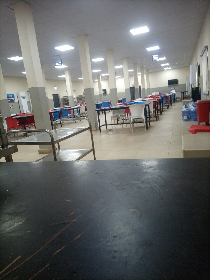 A long, bright cafeteria-style room is shown. Tables and chairs in blue and red are arranged in rows. White pillars line the room, supporting a ceiling with recessed lights. A metal cart is visible on the left. A dark surface occupies the foreground. A water cooler and other equipment are visible on the right.