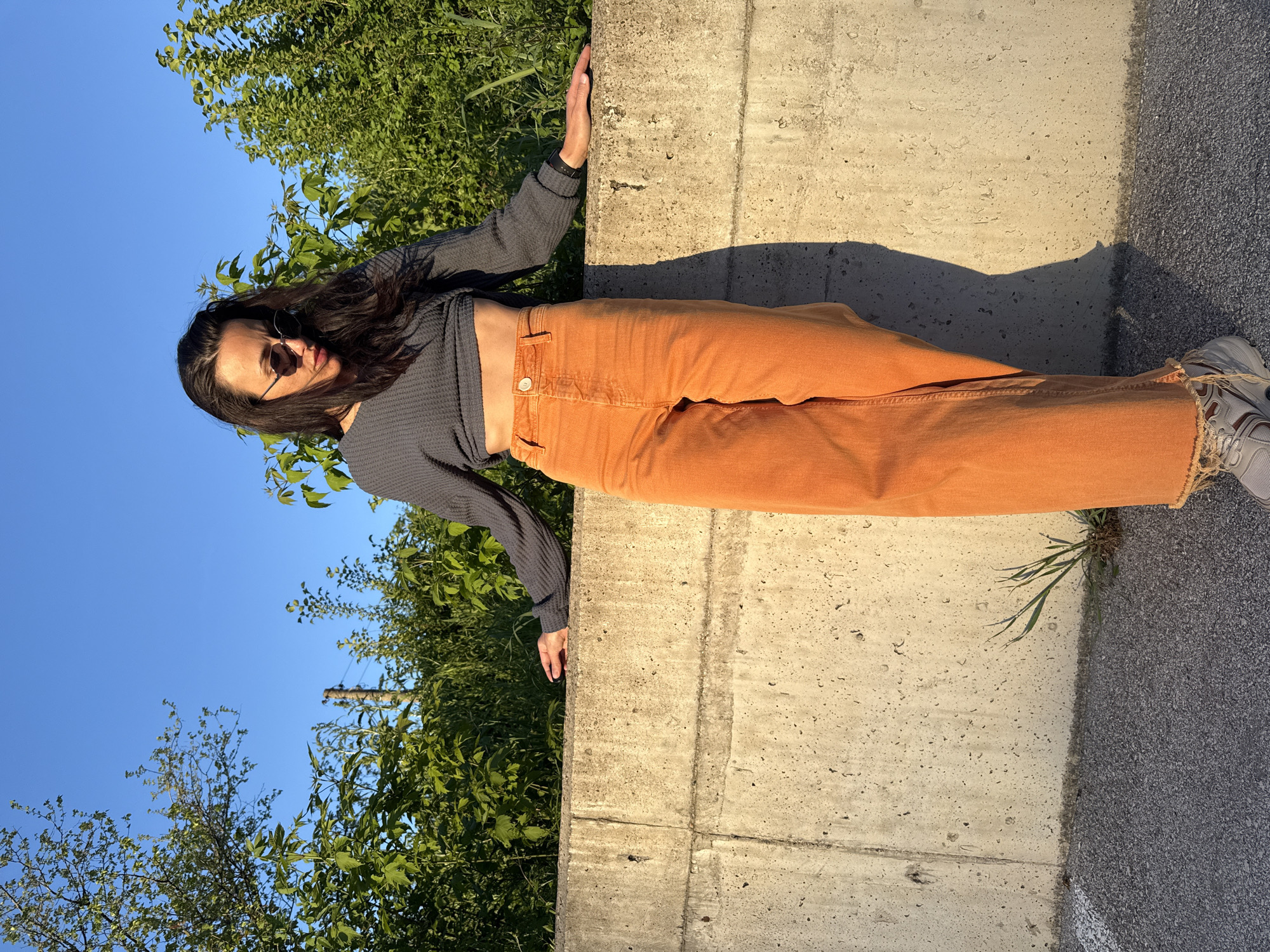 A woman with long dark hair and sunglasses leans against a concrete wall in an outdoor setting. She wears a dark grey long-sleeved shirt and orange pants. Her left hand rests on the wall to her left and the right foot can be seen near the bottom-right corner of the image. A shadow of a leg casts to the right side of the wall. Vegetation including grass and trees is visible behind the wall. A blue sky is above the trees. Asphalt can be seen near the wall where the right leg stands. The specific location within the city of Pleven, Bulgaria, cannot be determined from the image.