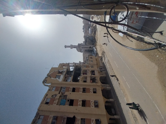 The image displays a street scene in Khan Yunis, Palestinian Territory, under a clear, bright sky with the sun low on the right, causing lens flare. A paved road with two visible lanes and white center markings stretches through the scene, flanked by dirt shoulders.
On the left, a multi-story, light-colored building is severely damaged, with large sections of its facade missing, exposing interior rooms and structural elements. Numerous smaller holes are visible on its remaining exterior walls. In the distant background, a mosque with a light dome and a tall, slender minaret stands amidst widespread destruction, characterized by extensive rubble and partially collapsed buildings.
Several people are present on the road: two individuals walk together on the left side in the foreground, casting long shadows; a solitary figure walks on the right side of the road in the mid-ground; and a group of three people walks further back in the distance, near the center of the road.
The immediate foreground on the right is dominated by a cluster of black electrical wires and cables, attached to an unseen pole or structure, partially obscuring a white banner with Arabic text and an orange symbol. The urban environment exhibits extensive damage and debris, particularly in the background.