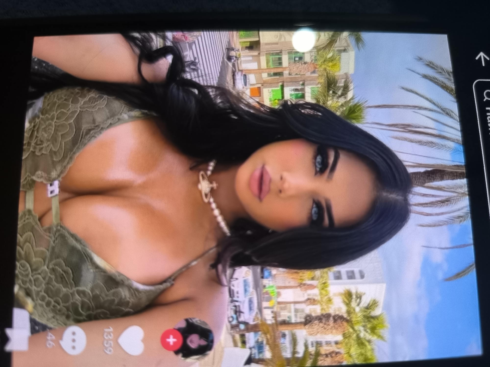 The image is a poorly lit, vertically oriented screenshot of a TikTok video. The video features a woman with dark hair and heavy makeup, wearing a green lace bra and a pearl necklace. She is posing with buildings and palm trees in the background. The user interface elements of TikTok are visible, including icons for comments, likes, and sharing. The image quality is low, with significant blur and pixelation. The location is stated to be in Bălți, Moldova, but there is no visual information within the image to support or refute this claim. The scene is casual and appears to be a selfie or a posed video for social media.