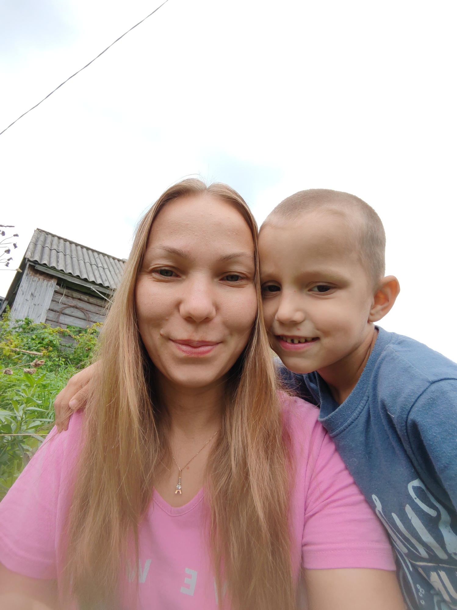 A medium shot selfie features a woman and a young boy outdoors in a rural setting. The woman, with long blonde hair and wearing a pink t-shirt, smiles at the camera, with her left arm around the boy. The boy, with short hair and wearing a blue t-shirt, smiles widely, leaning his head towards the woman. In the background, a weathered wooden building with a grey corrugated roof is visible, surrounded by green foliage. The sky above is overcast. The location is identified as the city of Чухлэм, Russia.