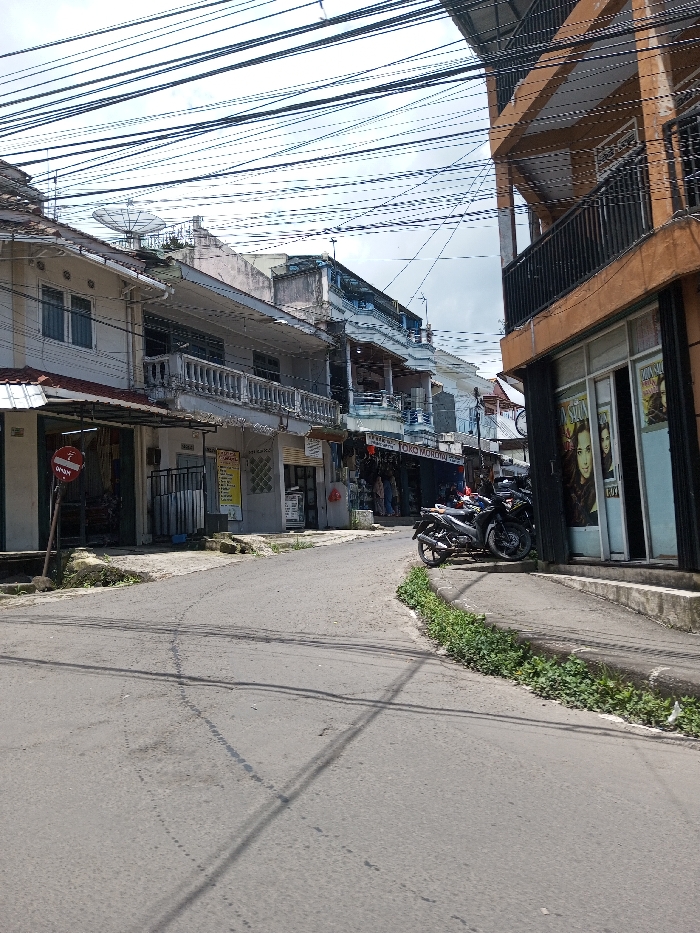 The image displays an urban street scene in Bagochen Boolingpang Ueling, Bhutan, under an overcast sky. A paved, slightly curved asphalt road, exhibiting signs of wear, occupies the foreground and midground. Buildings line both sides of the thoroughfare. On the left, a sequence of multi-story structures, primarily light-colored, features ground-level commercial fronts and upper-story residential sections with balconies and white balustrades. A satellite dish is visible on the roof of one building, and a red circular sign on a pole is positioned near the left curb. One visible sign on a shop reads "TOKO MORONOO." On the right, a distinct multi-story building with an orange-brown facade and black balcony railings features a ground-floor glass storefront displaying large posters depicting women's faces. Two dark-colored motorcycles are parked on the sidewalk adjacent to this building. Green foliage grows along the concrete curb on the right side of the road. A dense and complex network of black electrical and communication cables extends across the sky above the street and buildings. No individuals or specific human actions are clearly discernible within the frame.