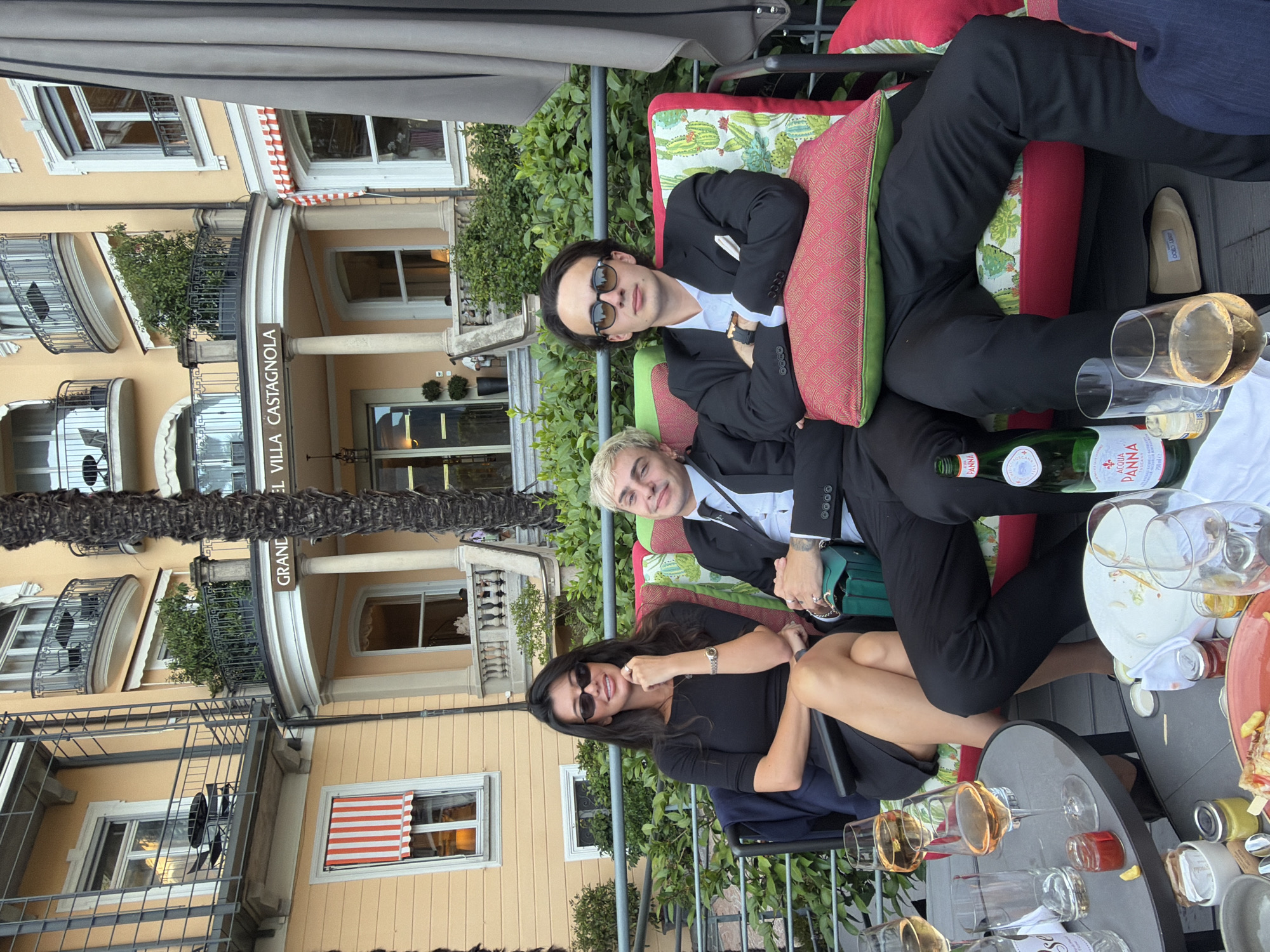 The media file shows a group of people, two men and one woman, seated at an outdoor table with drinks and plates of food. The man on the left is wearing a black suit with a white shirt and sunglasses. The man in the middle is wearing a similar suit. The woman on the right is wearing a black dress and sunglasses. They are seated on a bench with colorful cushions, in front of a building with balconies and windows. The building appears to be old, with architectural details suggesting a European setting. There is also greenery and foliage surrounding the area. On the table, there are wine glasses, a bottle of Acqua Panna water, and various plates. The overall scene suggests a relaxed social gathering, possibly a meal or drinks outdoors. The lighting suggests it is daytime, and the weather appears pleasant. The visible text reads "ACQUA PANNA" on the water bottle and "GRAND HOTEL VILLA CASTAGNOLA" on a sign on the building. The people's expressions and relaxed postures suggest they are enjoying themselves.