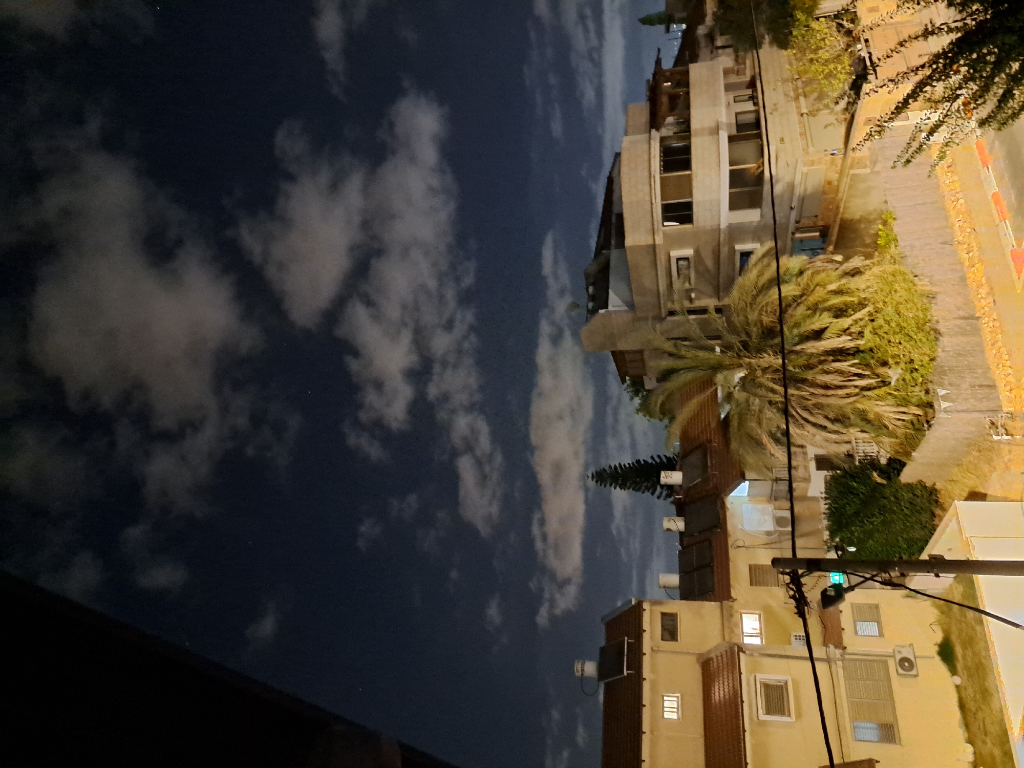 The image captures a night view of a residential area in Ramat HaSharon, Israel. The sky is dark blue with scattered white clouds, and a few faint stars are visible. In the foreground, a portion of a dark structure, possibly a balcony or roof overhang, is visible on the left.
The middle ground of the image features several buildings. On the left is a two-story residential building with a red-tiled roof. It is painted a light yellow and has windows with white frames. The building on the right is larger and more modern, with multiple levels and large windows. It is also painted in a light, neutral color. A tall, conical evergreen tree stands between these buildings. A large palm tree with dense fronds is located in front of the building on the right.
Below the buildings, a fence is visible, constructed from wood or similar material. Streetlights and power lines are also present, with a streetlamp prominently displayed on a pole in the lower-middle section of the image. Some vegetation, including bushes and trees, can be seen around the buildings and fence. The ground level appears to be a street or pathway, with some light colored and red markings indicating a curb or road edge. The scene suggests a peaceful evening in a suburban or urban residential neighborhood.