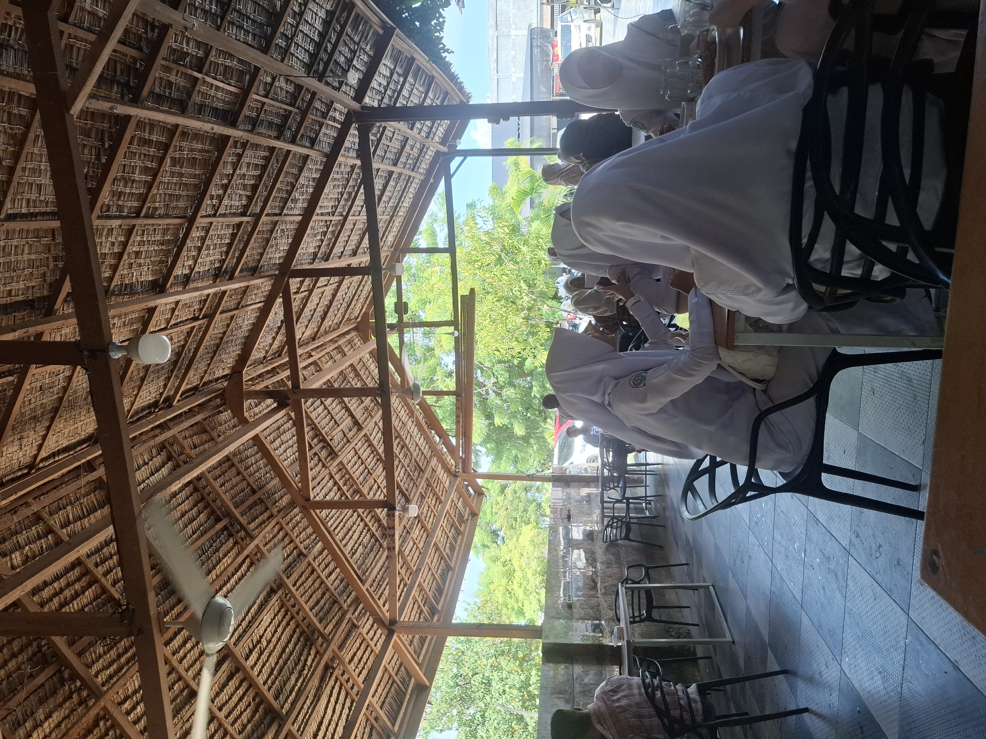 The image depicts an outdoor seating area of a restaurant in Keuniree, Indonesia. The area is covered by a thatched roof supported by wooden beams, with a ceiling fan visible. Several people, predominantly young women wearing hijabs and white uniforms, are seated at tables, engaged in conversation and dining. The floor is tiled, and in the background, there are stone walls, trees, and a few parked vehicles. The overall atmosphere suggests a casual dining or break setting.