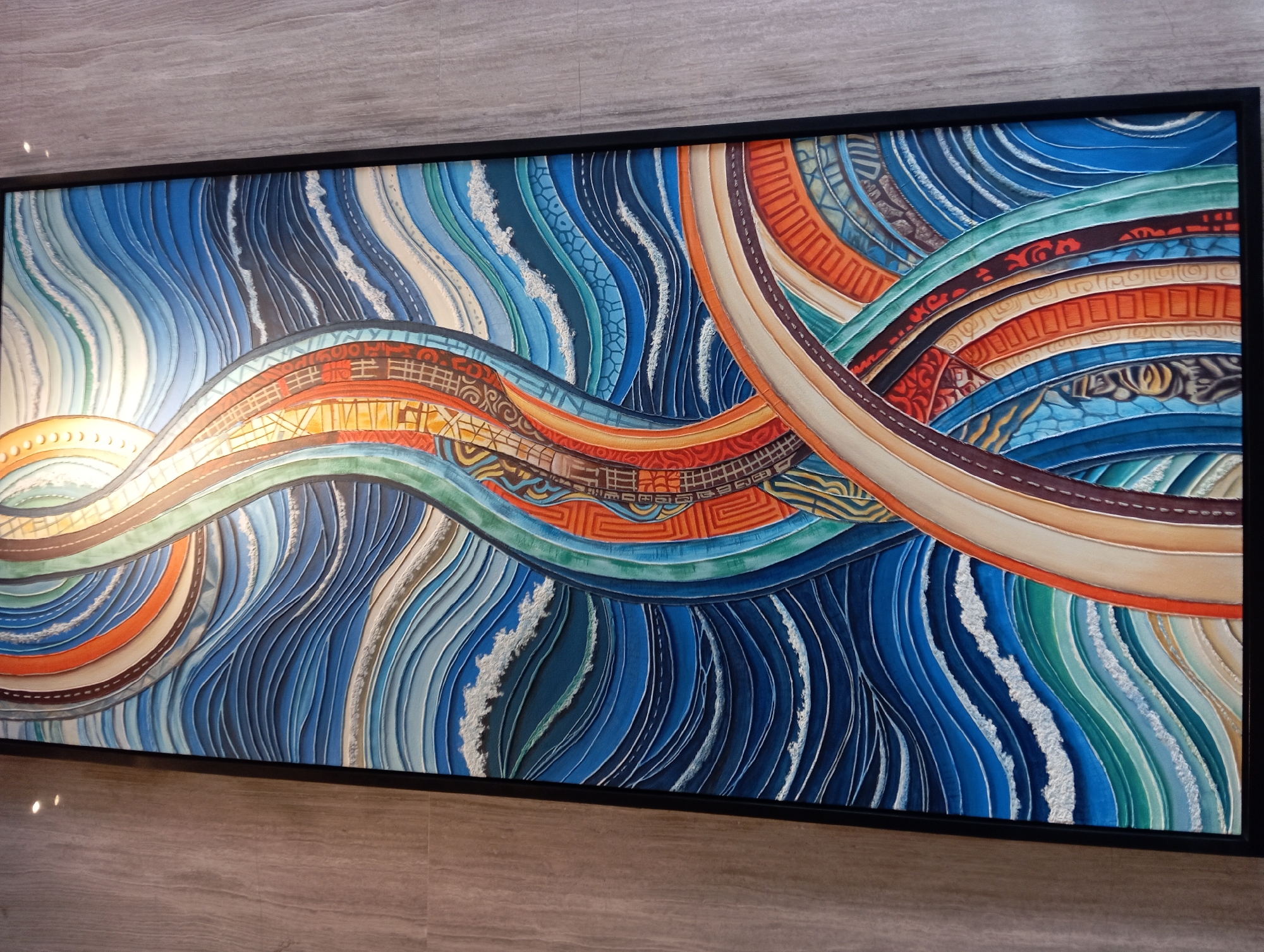 The image shows a vertical, framed artwork mounted on a wall with a grey, textured surface. The artwork itself is a vibrant, abstract painting with a dominant color palette of blues, oranges, and greens, featuring curved lines and intricate patterns. The composition appears to depict stylized waves or a flowing pathway. The painting is located in the lobby of a Starbucks store in Nha Trang, Vietnam. The wall texture suggests a modern interior design, and the lighting on the wall indicates indoor illumination. There are no people visible in the image.