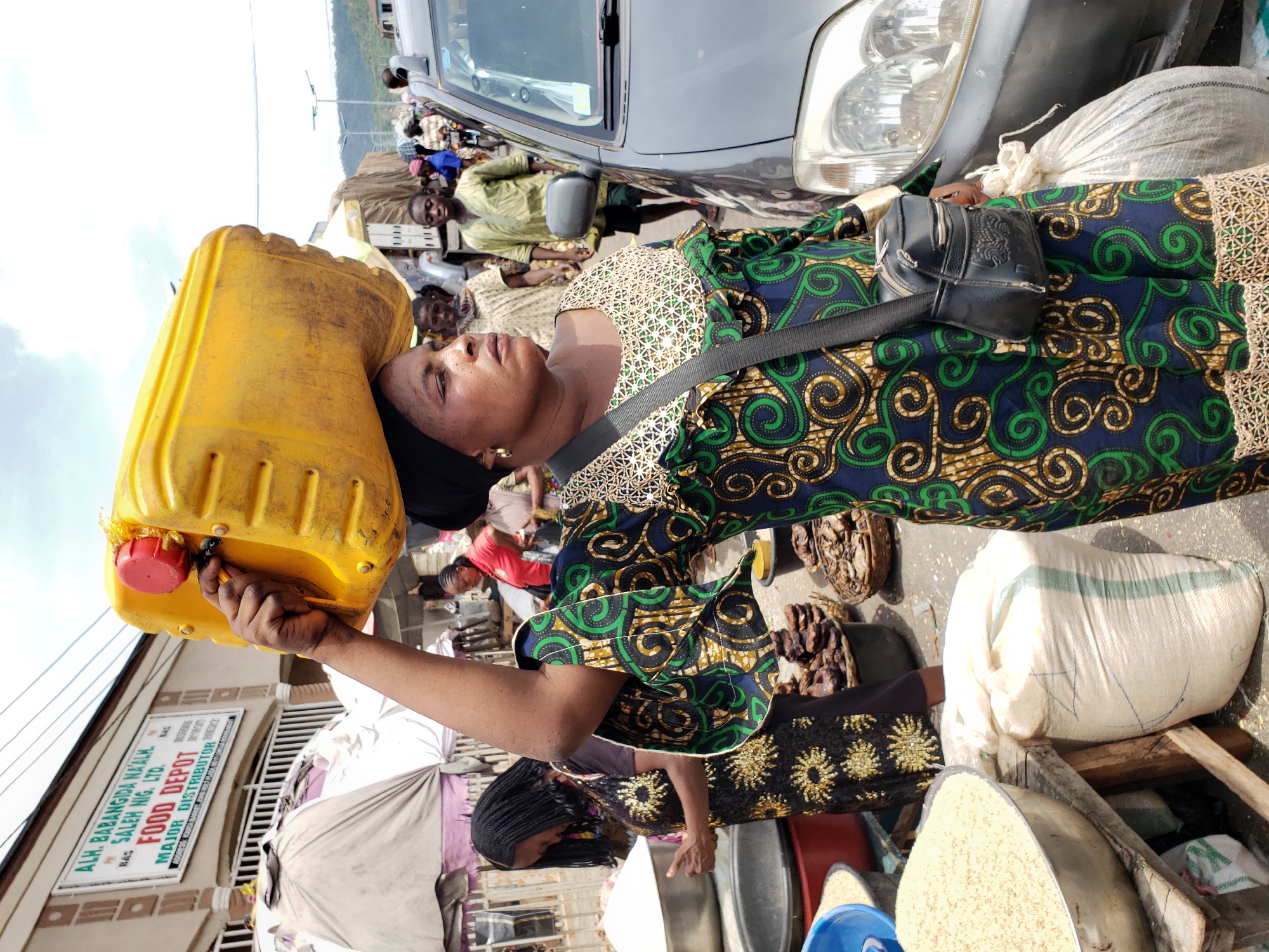 The image depicts a woman in Lokoja, Nigeria, balancing a yellow plastic jerrycan on her head. She is wearing a patterned green, blue, and gold dress with ruffled sleeves and a black shoulder bag. The scene is a crowded market setting. Other people are visible in the background, along with goods for sale, including bags of what appears to be rice. In the background, one can see the front of a building that contains the sign "ALH. BABANGIDA NAALH. SALEH NIG. LTD. FOOD DEPOT MAJOR DISTRIBUTOR". A gray vehicle is also present.