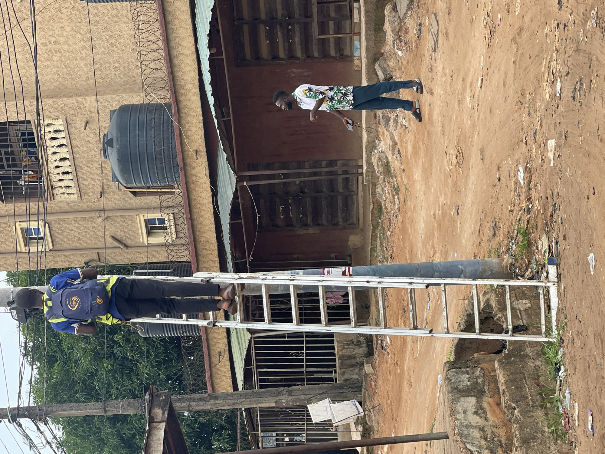 In Nkpor, Nigeria, a man in a blue shirt, black pants, and a blue and yellow backpack is climbing a silver aluminum ladder leaning against a utility pole. Another man in a white, floral-patterned shirt and dark pants stands on a dirt path next to a building. Overhead electrical wires are visible, connecting to the pole. A black water tank is mounted on the side of a building with barred windows and barbed wire along the roofline. The foreground shows a cluttered dirt area with litter.