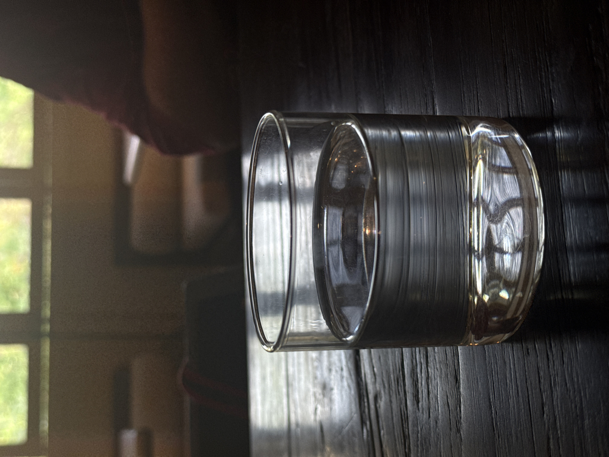 The image, when viewed upright, displays a clear, double-walled glass tumbler resting on a dark, textured wooden surface, likely a table. The glass contains a small amount of transparent liquid, possibly water, at its base. Bright natural light illuminates the scene, creating strong reflections on the glass and the glossy wood grain of the table.
In the background, a large window with multiple panes is visible, framing a vibrant green outdoor scene, indicative of lush foliage or a garden. The illumination suggests it is daytime. The glass itself captures and distorts reflections of the window frames and surrounding environment.
In the upper left corner of the image (when upright), a soft, blurred shape with reddish-brown tones can be seen, which might be a person's arm, hand, or draped clothing, though no distinct human features are identifiable. The scene appears still, with no specific activity taking place. There is no visible text in the image.
The location context for this media file is North Kuta, Indonesia.