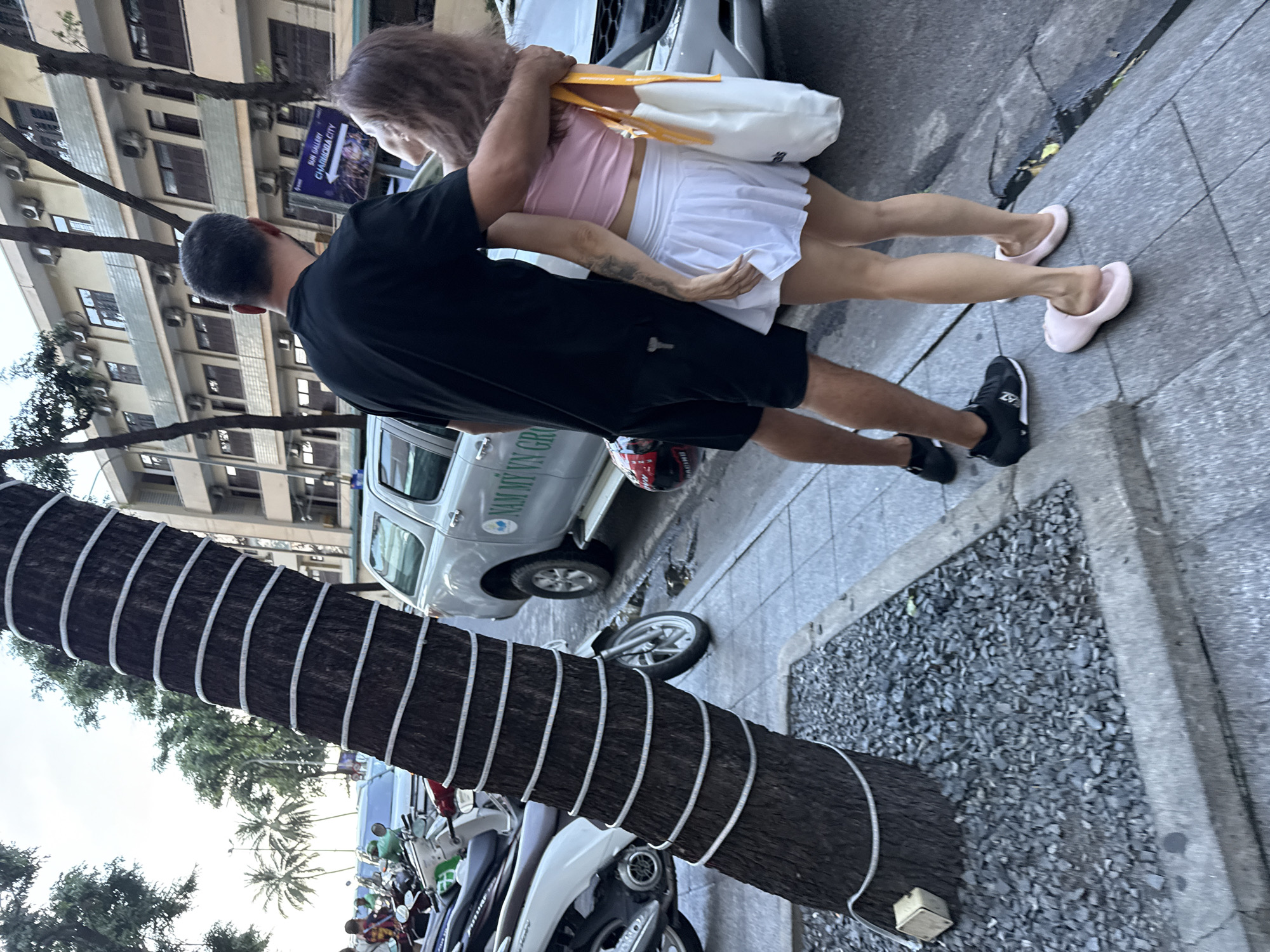 The image depicts a street scene in Nha Trang Ward, Vietnam. In the foreground, a man with short, dark hair embraces a woman from behind. The man is wearing a black t-shirt and black shorts, while the woman is dressed in a pink crop top and a white pleated skirt. She is carrying a white tote bag with yellow straps. Their lower legs and feet are visible, with the man wearing black sneakers and the woman wearing pink slides.
To their left, a large tree trunk is wrapped with white rope lights. Behind them, parked motorcycles are visible, along with a silver pick-up truck. The truck has text on its side that reads "NAM MY VN GRO...". A street sign with an arrow is also present, displaying the text "SUN GALLERY CHAUDRA CITY". The background features a multi-story building with numerous windows and air conditioning units, typical of urban architecture. The ground is paved with square tiles, and there are patches of gravel around the base of the tree. Palm trees are visible in the distance, suggesting a tropical climate. The overall impression is an everyday street scene in a Vietnamese city.