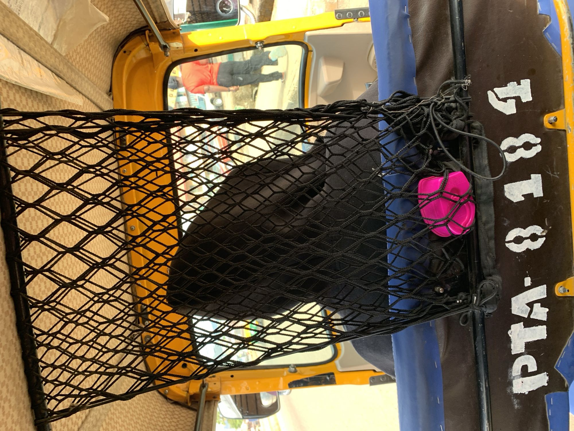 The image depicts the interior of a yellow vehicle, possibly a taxi, with a black net attached to the front seat and roof. A dark-colored bag is secured within the net, along with a pink circular container. The passenger seat is blue. Visible through the windows are reflections of people. The vehicle's license plate reads "PTA - 8184". There is no verifiable indication of it being located in Rafin Par, Nigeria.