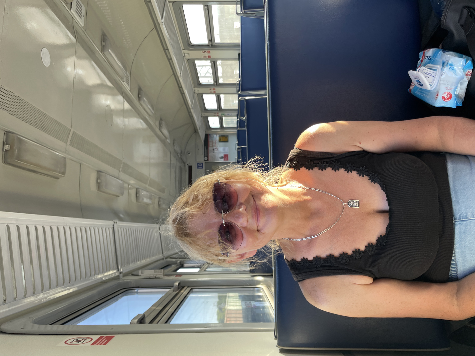 The media file shows a woman with blonde hair and sunglasses posing for a selfie inside a train compartment. She is wearing a black top with a floral trim and a silver necklace with a pendant. The train interior is visible, with rows of blue seats and windows on the left side. Sunlight streams through the windows, suggesting it is daytime. A packet of wet wipes is visible on the seat next to her. The background shows the interior of a train car, with windows and seats. The image is oriented vertically, with the woman's head at the bottom and the train interior extending upwards. There are no visible signs of weather conditions. The setting appears to be a train in Artyshta, Russia.