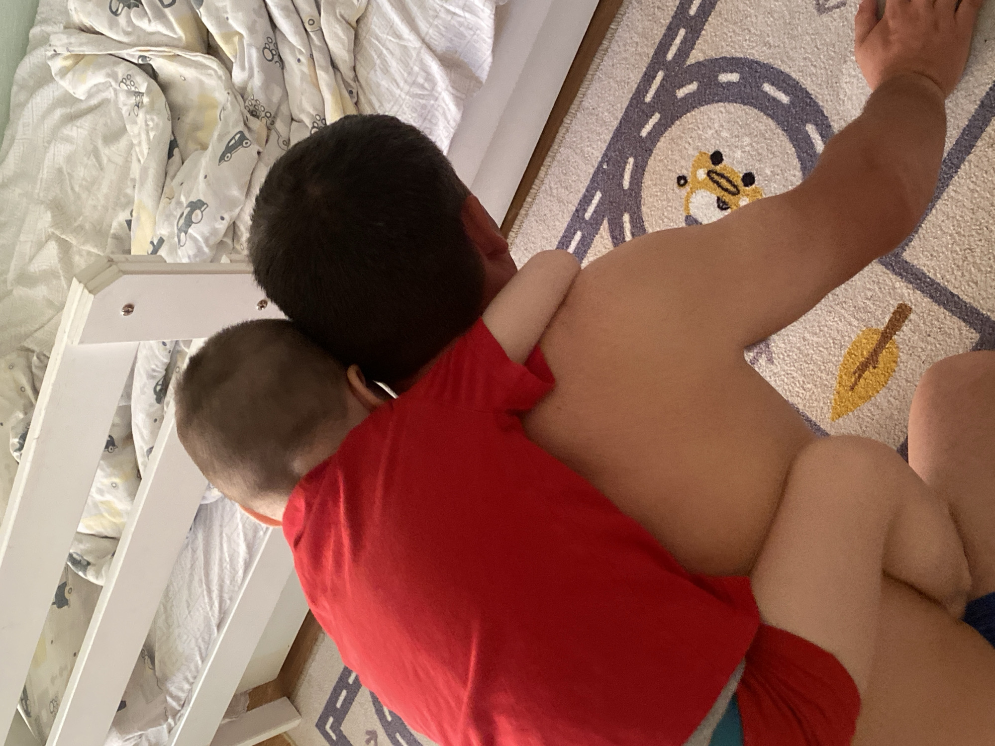 A photo shows a child in a red shirt clinging to a man's back. They are indoors, near a crib with white rails and patterned bedding. The floor is covered with a rug depicting a road with cars. The location is not evident from the image.