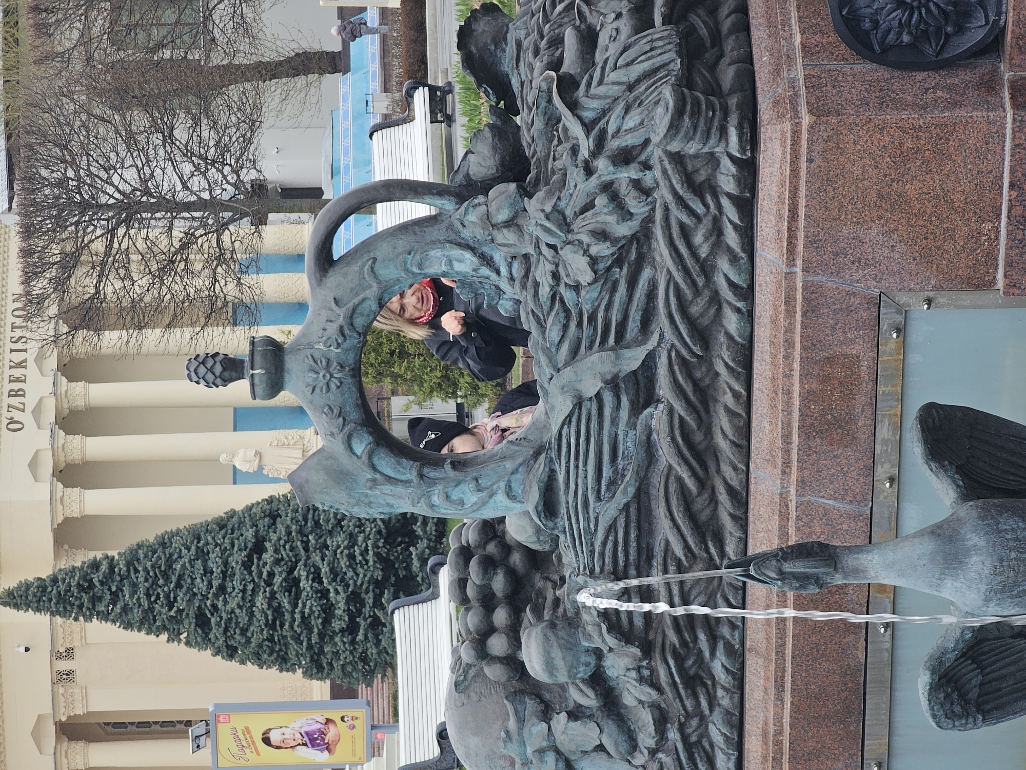 The image shows a bronze fountain sculpture of two swans spewing water in Moscow, Russia. Two people, a woman with a red scarf and a person in a black beanie, are posing for a photo through the sculpture. The building in the background is labeled "O'zbekiston". A sign in the foreground features a child holding a gift. A green pine tree is located next to the sign.