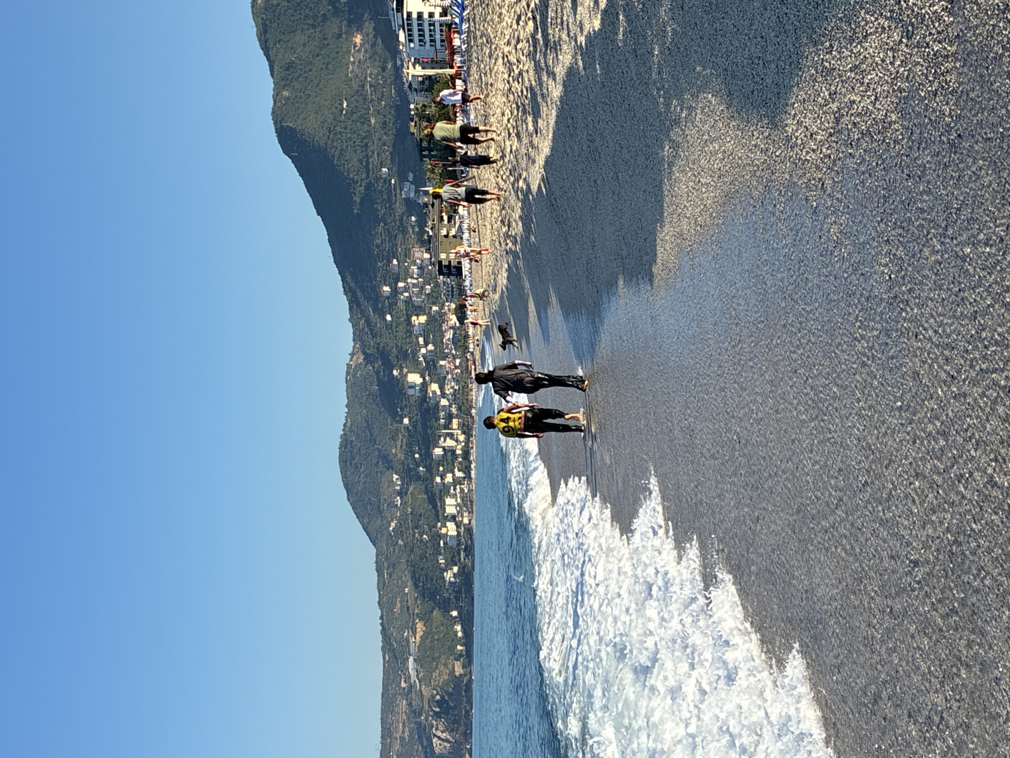 The image depicts a beachfront scene in Alanya, Turkey, with a clear blue sky. A wide, pebbly beach is in the foreground, with the gentle waves of the Mediterranean Sea lapping at the shore. In the middle ground, two individuals are walking away from the viewer along the water's edge. One person wears a yellow and black patterned shirt and dark pants, while the other wears dark clothing. Further up the beach, several other people are visible, some walking, some with dogs, and a group of people relaxing on beach chairs under umbrellas in the distance. A town with multi-story buildings and lush green hillsides forms the backdrop, suggesting a coastal urban environment. The light indicates it is likely late afternoon or early morning, casting long shadows across the sand.