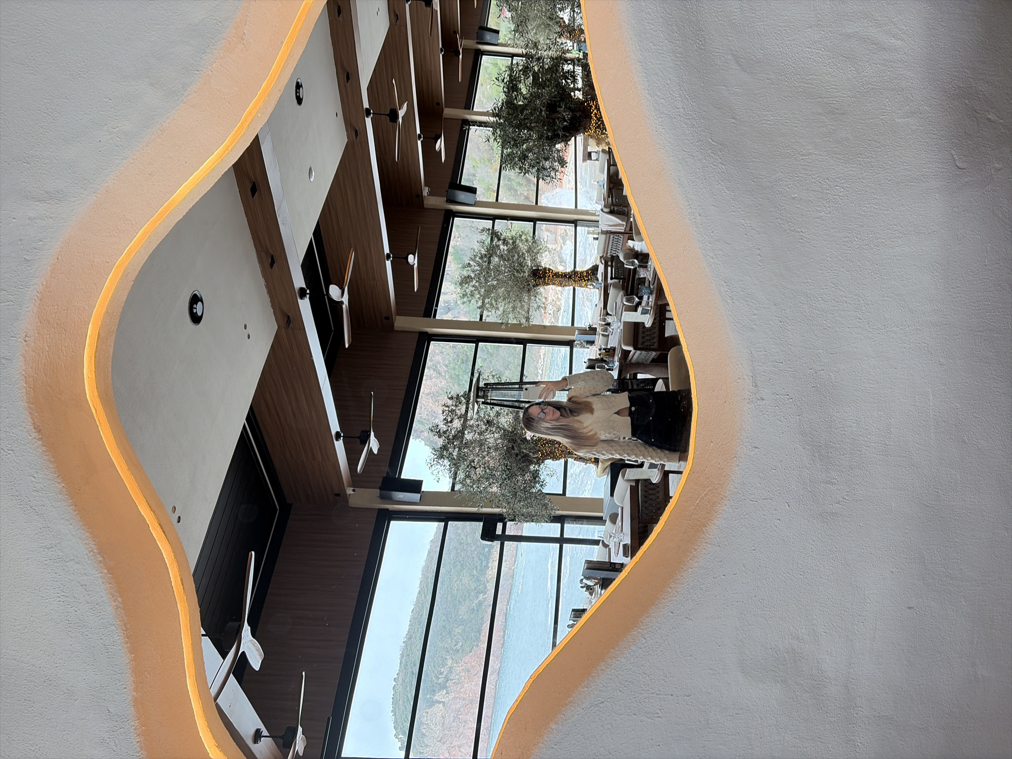 A female individual is captured in a selfie reflected within a large, irregularly shaped wall mirror, located inside a restaurant or dining establishment in the city of Unknown, Unknown. The mirror's frame is textured white with an illuminated orange-yellow strip along its inner edge. The individual, appearing to be a young adult with light-colored, long hair and glasses, is holding a white smartphone up to take the photograph. She is dressed in a cream-colored, long-sleeved knitted top and dark bottoms.
The restaurant interior features a contemporary design with a mixture of dark wood paneling and lighter sections on the ceiling, equipped with multiple ceiling fans and recessed lighting fixtures. Several dining tables, set with glassware and crockery, are visible in the background, along with upholstered chairs. Large, floor-to-ceiling windows line the exterior wall, offering a panoramic view of a body of water, likely a sea or large lake, bordered by lush, green, and rocky hills or mountains under an overcast sky. Several potted trees, some adorned with small fairy lights, are positioned near the windows, enhancing the interior decor. A tall, dark outdoor patio heater is also visible near one of the trees. No direct interactions between individuals are discernible, only the solitary action of the individual taking a selfie.