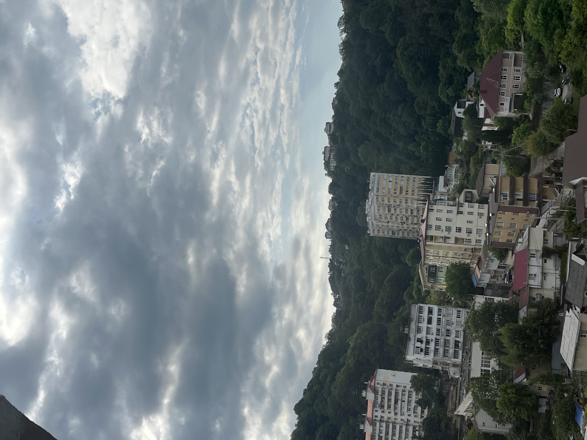 The image captures a view of a city nestled in a hilly, densely forested area under a cloudy sky. Several multi-story apartment buildings and houses are visible, some with red-tiled roofs. The buildings are clustered together, with roads and smaller structures interspersed. The forest covers the hillsides, with trees extending down towards the buildings. The sky is filled with dark, heavy clouds, with some patches of lighter sky and sunlight breaking through. The overall impression is of an urban settlement integrated into a natural, somewhat rugged landscape, likely in a coastal or mountainous region. The presence of numerous apartment blocks suggests a developed urban area. The architecture of the buildings, particularly the tall apartment complexes with numerous balconies, hints at a post-Soviet era construction style, common in many Russian cities, including Sochi. The lush greenery of the forested hills suggests a temperate or subtropical climate, which is characteristic of Sochi.