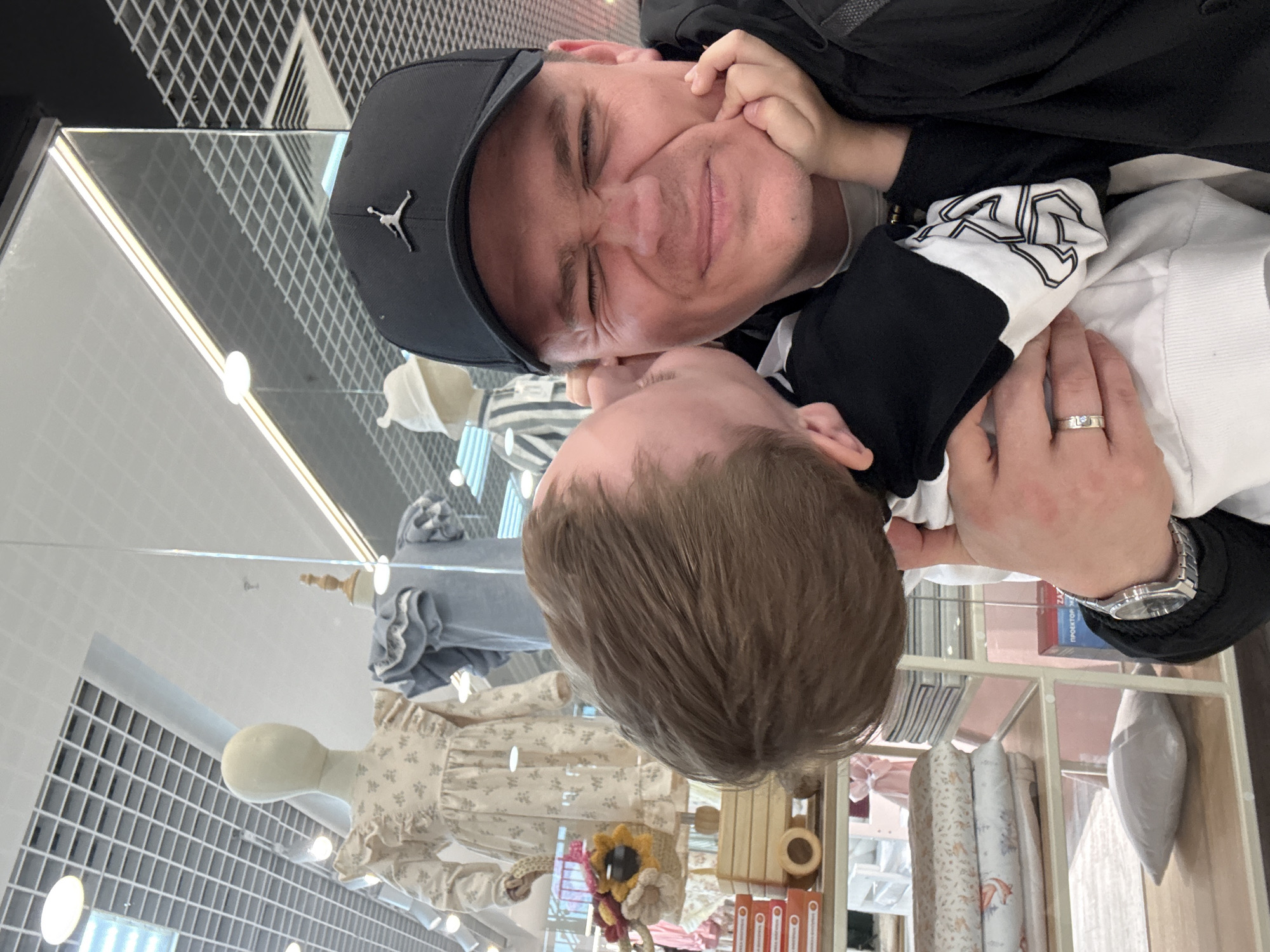 An adult male is depicted embracing a young child in a retail store setting in Vladivostok, Russia. The man, positioned on the right, looks directly at the camera with his eyes scrunched closed and his face contorted in a playful expression. He wears a black baseball cap with a silver Jordan 'Jumpman' logo, a black jacket over a white shirt, a silver-toned watch on his left wrist, and a silver ring with a light blue stone on his left ring finger. The child, partially visible on the left, has light brown hair and is pressing its cheek against the man's face, appearing to be giving a kiss or hug. The child wears a black and white long-sleeved top with a distinctive pattern on the white sleeve.
The background, visible through a glass partition, reveals a children's clothing or goods store. Three child-sized mannequins are present: one wears a cream-colored dress with a floral pattern and ruffled details, another wears a light blue ruffled top, and a third wears a white and blue striped top with a white hat. A shelving unit displays folded patterned textiles, a small white pillow, and a wooden toy wagon containing knitted floral decorations. A red box with Cyrillic text, reading "ПРОЕКТОР ЖИЗНИ" (Projector of Life), is visible on one of the shelves. The ceiling above features a grid pattern with embedded circular and linear lighting fixtures.