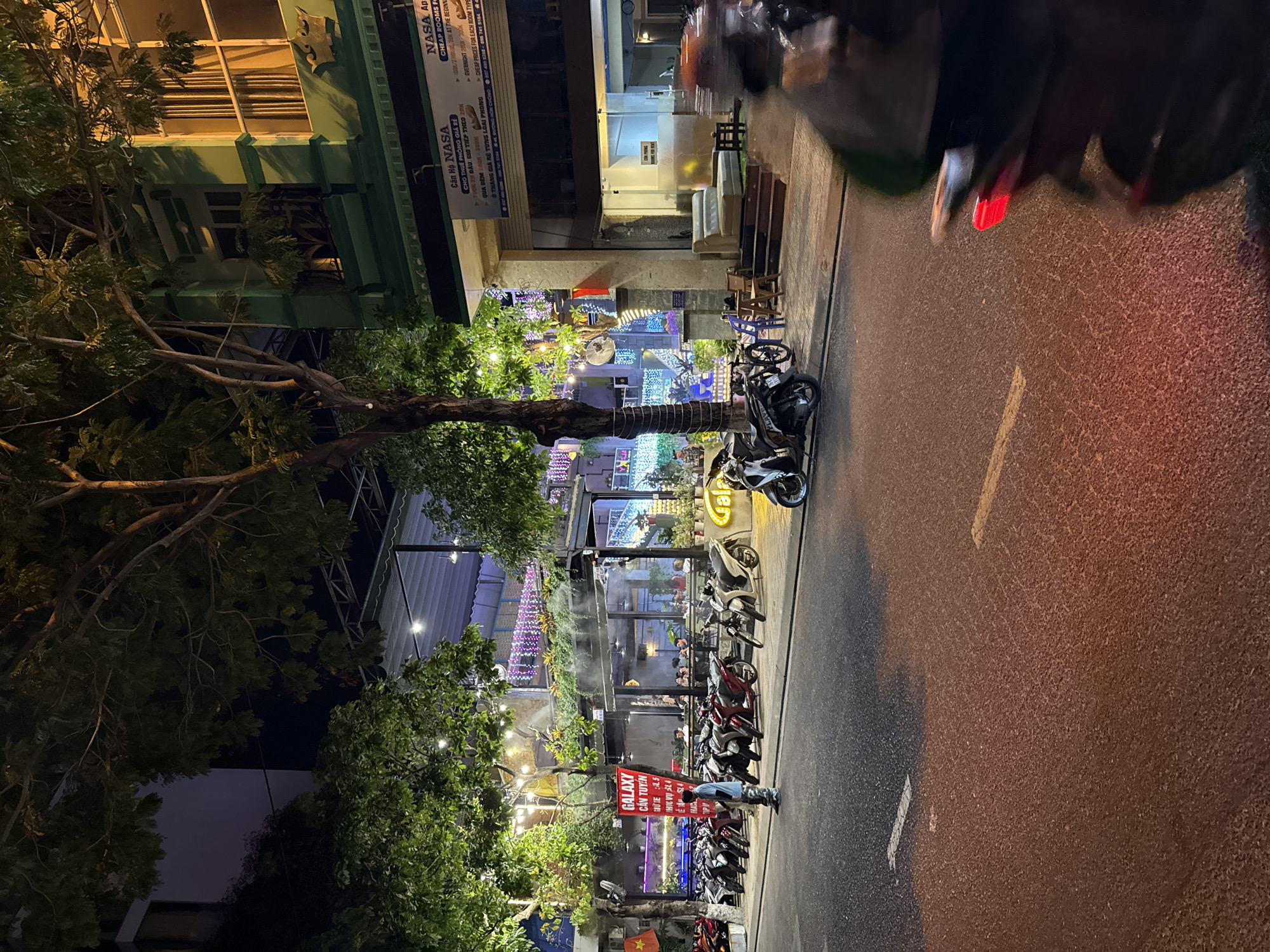 This is a night-time exterior shot of a street scene in Bac Nha Trang Ward, Vietnam. A row of parked motorcycles lines the sidewalk in front of a building with a green facade. Above the motorcycles, a canopy shades an outdoor seating area of a restaurant or cafe, decorated with string lights and potted plants. The building itself appears to be multi-story, with windows visible on the upper floors. Signs for "GALAXY" and "NASA" are present. A person stands on the sidewalk next to the motorcycles. A motorcycle in the foreground is blurred, indicating movement. The street is paved with asphalt, with visible lane markings. The overall atmosphere is that of a lively urban area at night, with commercial activity and transportation.