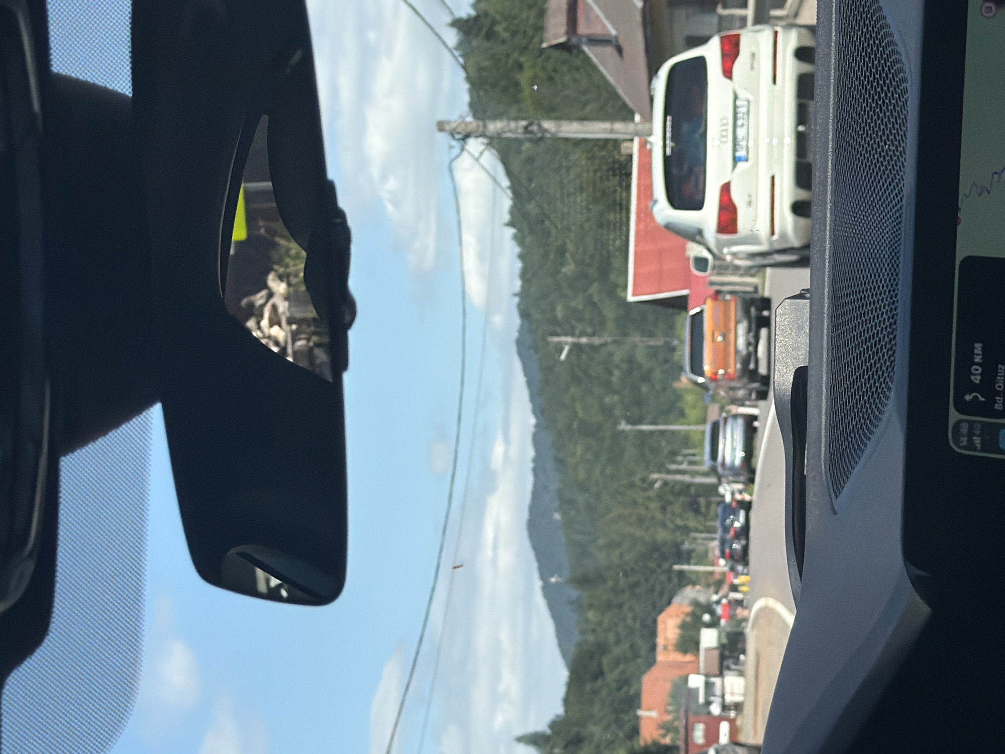 The image depicts a view from inside a vehicle, looking forward through the windshield and at the interior rearview mirror. Traffic is visible on a road lined with houses and lush green hills in the background. The sky is blue with scattered clouds. Several cars are stopped or moving slowly in traffic, including a white Audi SUV in the foreground, an orange pickup truck behind it, and other vehicles further down the road. A power line runs horizontally across the middle of the frame, with a utility pole visible on the right. The scene appears to be in a semi-rural area. The navigation display on the car's dashboard shows "40 KM" and "Bd. Oituz," indicating the location is on Boulevard Oituz in Romania.