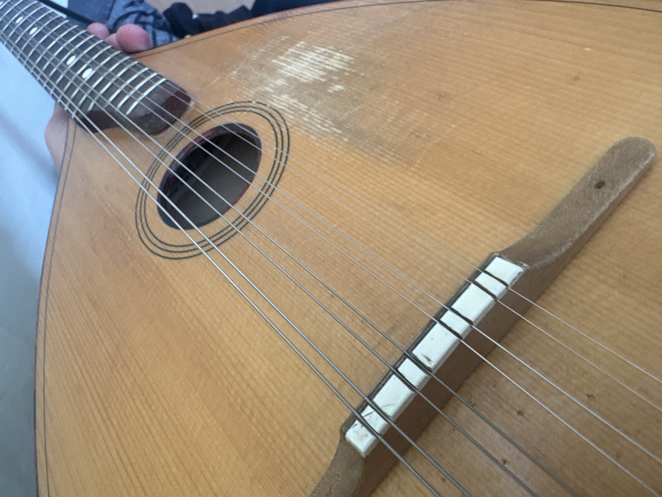 A close-up, angled view depicts an eight-stringed musical instrument, consistent with a mandolin or domra, being held indoors in Kotel, Bulgaria. The instrument's light-toned wooden soundboard displays vertical grain patterns and extensive abrasion with scratches below the central round soundhole. The soundhole features concentric black decorative rings. Eight metallic strings are arranged in four courses, extending across a wooden bridge with a segmented white saddle, which is affixed to the soundboard. A human hand grips a portion of the instrument's dark fretboard, revealing visible frets. The background is a light blue-grey wall.