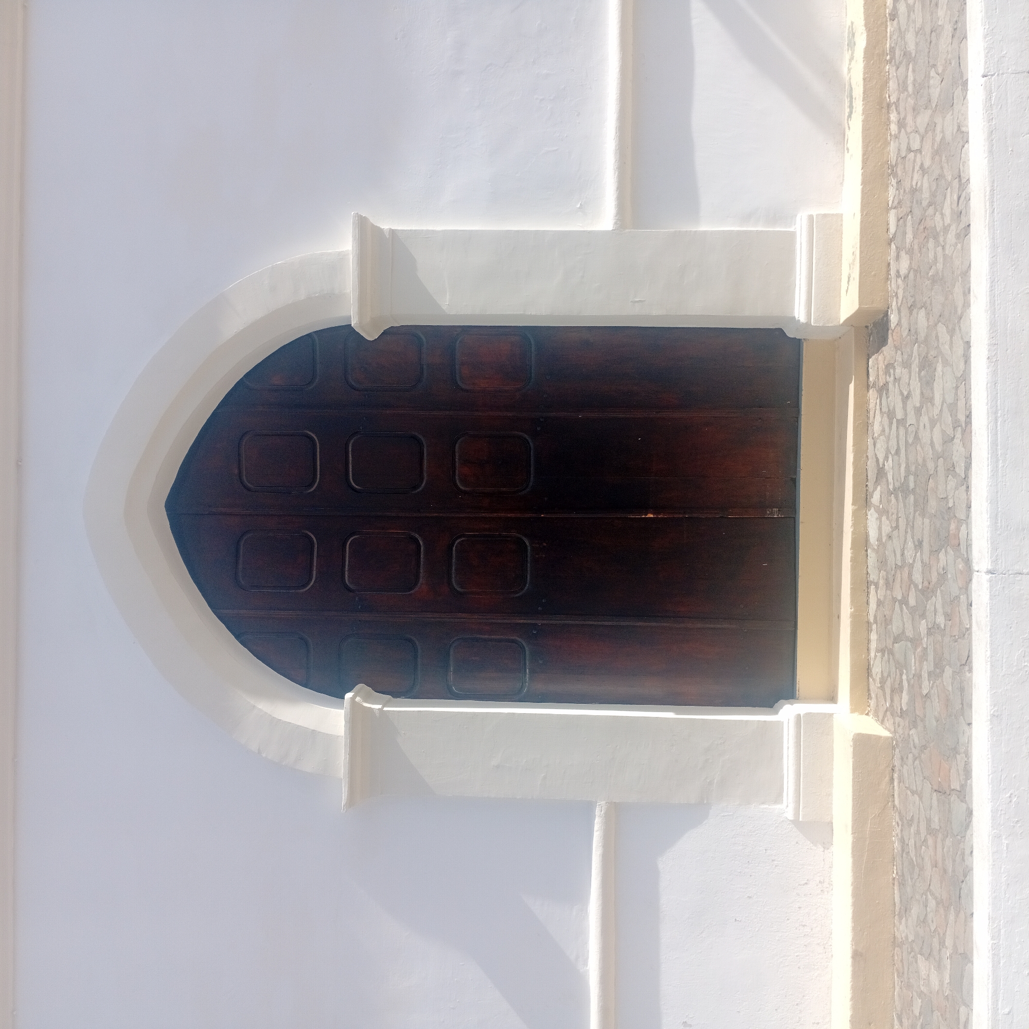 A dark wooden door with a pointed arch is set into a white wall. The door is divided into rectangular panels. The arch is framed with a white trim, and the wall has decorative molding and stonework to the right. The location is unknown, Unknown.
