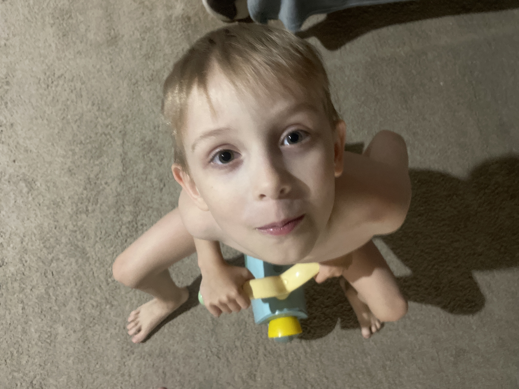 A high-angle shot depicts a young, fair-skinned boy indoors. The boy has blonde hair, light eyes, and is holding a toy scooter. His torso is visible, but his clothing is not apparent. The toy scooter is light blue with a yellow handle and base. The background shows a neutral-toned carpet. The location is not discernible in the image, and there's no basis to indicate it's Baranovka, Russia.