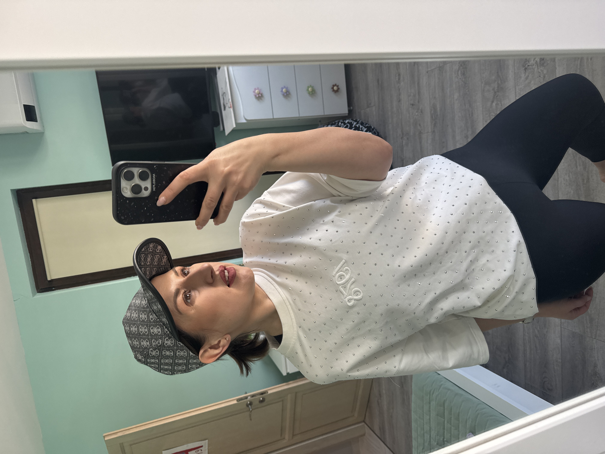 A woman is taking a selfie in a mirror. She is wearing a patterned baseball cap, a white shirt with silver studs, and black leggings. She is holding a black phone in her right hand. The reflection in the mirror shows a room with mint-green walls, a cabinet with decorative flower knobs, a black framed flat screen TV, and light gray flooring. A sign is visible on the lower left corner of the photo. The location is inside a building, likely an apartment, in Sofia, Bulgaria.