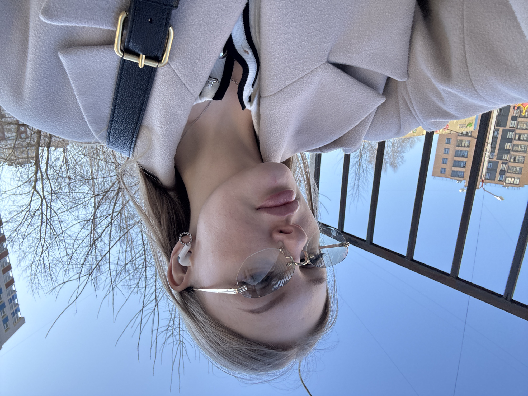 A young woman in Perm, Russia, takes a selfie outdoors. She is wearing a light-colored coat, sunglasses, and a small earring. A black purse strap crosses her torso. In the background, bare trees, buildings, and a clear blue sky can be seen. The image is taken at an unusual angle, with the woman's face oriented towards the bottom of the frame. Metal bars are visible in the foreground.