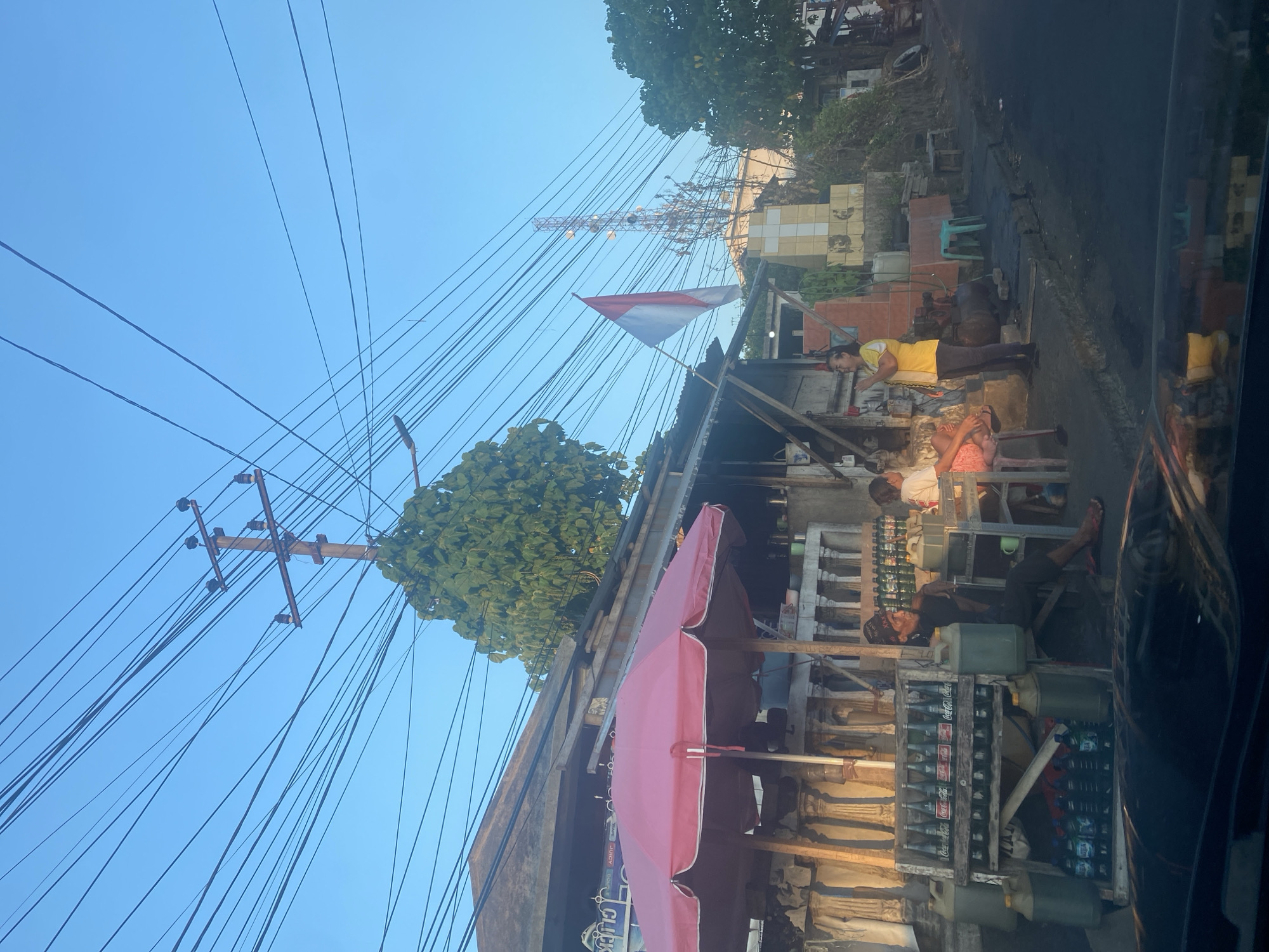 The image depicts a street scene in Manado, Indonesia, with a clear blue sky overhead. Numerous power lines crisscross the sky, originating from a utility pole in the mid-ground. In the foreground, a roadside stall is set up under a large pink umbrella, selling various bottled drinks, including Coca-Cola, and larger containers of what appear to be fuel or water.
Several individuals are present at the stall. A man with a dark hat sits on a chair, looking to his left. Two other people are seated at a table, possibly customers. A woman in a yellow shirt and dark pants stands near the stall, gesturing with her hands as if in conversation.
To the right of the stall, a small monument with a cross shape is visible. Further in the background, a tall telecommunications tower stands against the sky, and dense foliage from trees can be seen. The overall setting suggests a modest urban environment, characteristic of many street-side businesses in the city. The road surface appears to be asphalt.
