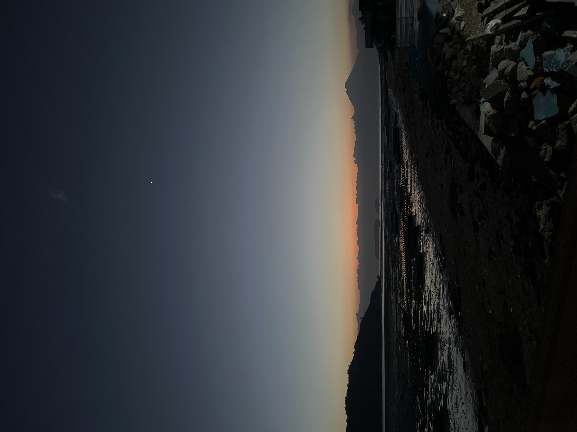A twilight scene unfolds over a coastal landscape in Kertasari, Indonesia, featuring a beach, the sea, and distant landforms. The sky transitions from a deep indigo at the top to a soft gradient of blue and orange near the horizon, suggesting the period just before sunrise or after sunset. Two bright celestial bodies, likely planets or bright stars, are visible in the upper portion of the sky, along with a wispy cloud formation.
The foreground is dominated by a dark, sandy beach littered with rocks and debris. To the left, a muddy, exposed seabed is visible, reflecting the dim light and dotted with what appear to be remnants of structures or boats. A dark, silhouetted hill or island rises from the left side of the frame, its outline sharp against the fading sky.
Across the water, a hazy mountain range stretches horizontally, with a distinct, conical peak standing out on the right side. The water itself is calm, with the faint glow of the horizon reflecting on its surface. On the far right of the image, the dark silhouettes of buildings and construction materials are visible, indicating some level of human habitation or development near the coast. The overall atmosphere is serene and tranquil, characteristic of dawn or dusk. There are no people or animals visible in the frame.