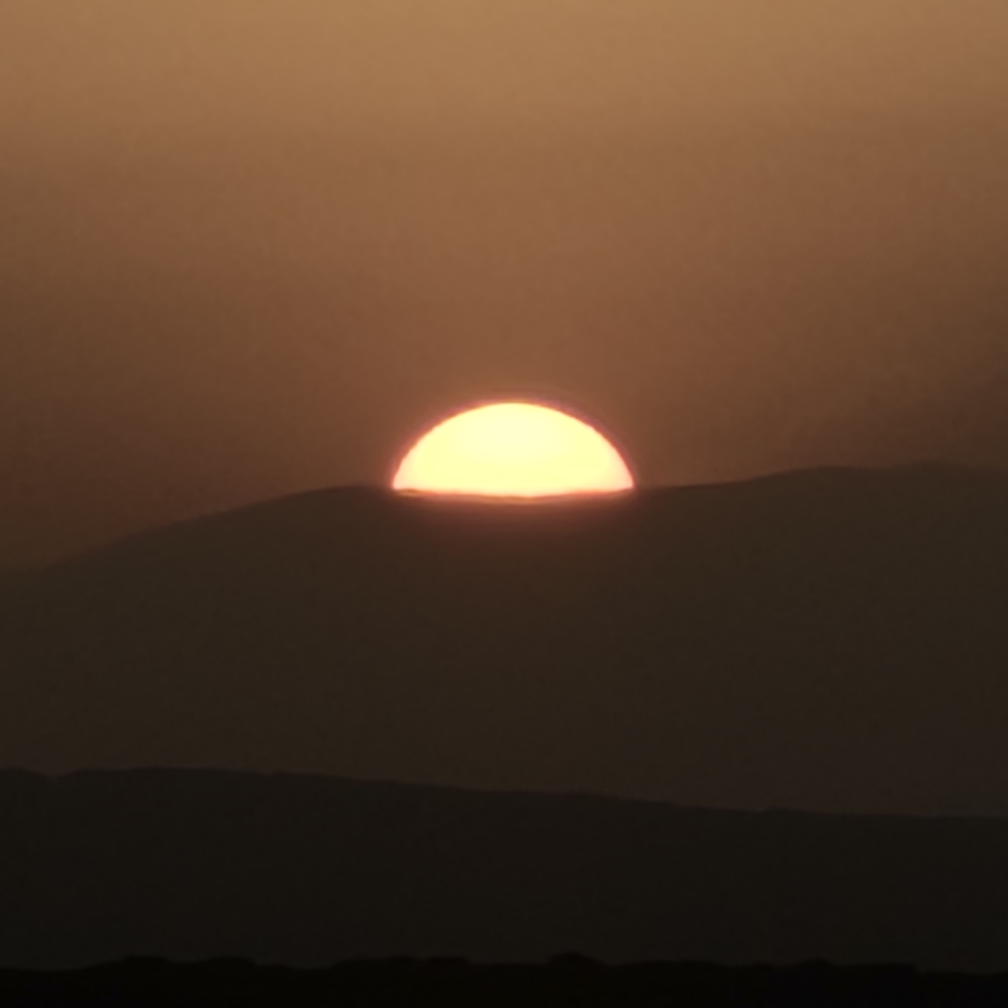 The image shows a sunset over a silhouetted mountainous landscape. The sun is a bright, glowing semi-circle just above the horizon, casting a warm, orange light that fills the sky. The landscape below is dark and indistinct, with layers of shadow suggesting hills or mountains. The sky is a gradient of warm oranges and browns, fading to a darker shade at the top of the frame. The overall impression is one of natural beauty and tranquility, as the day draws to a close. The location is specified as Algora, Spain, indicating a rural or natural setting within that region. There are no people or specific objects visible that would provide further context or action within the scene.