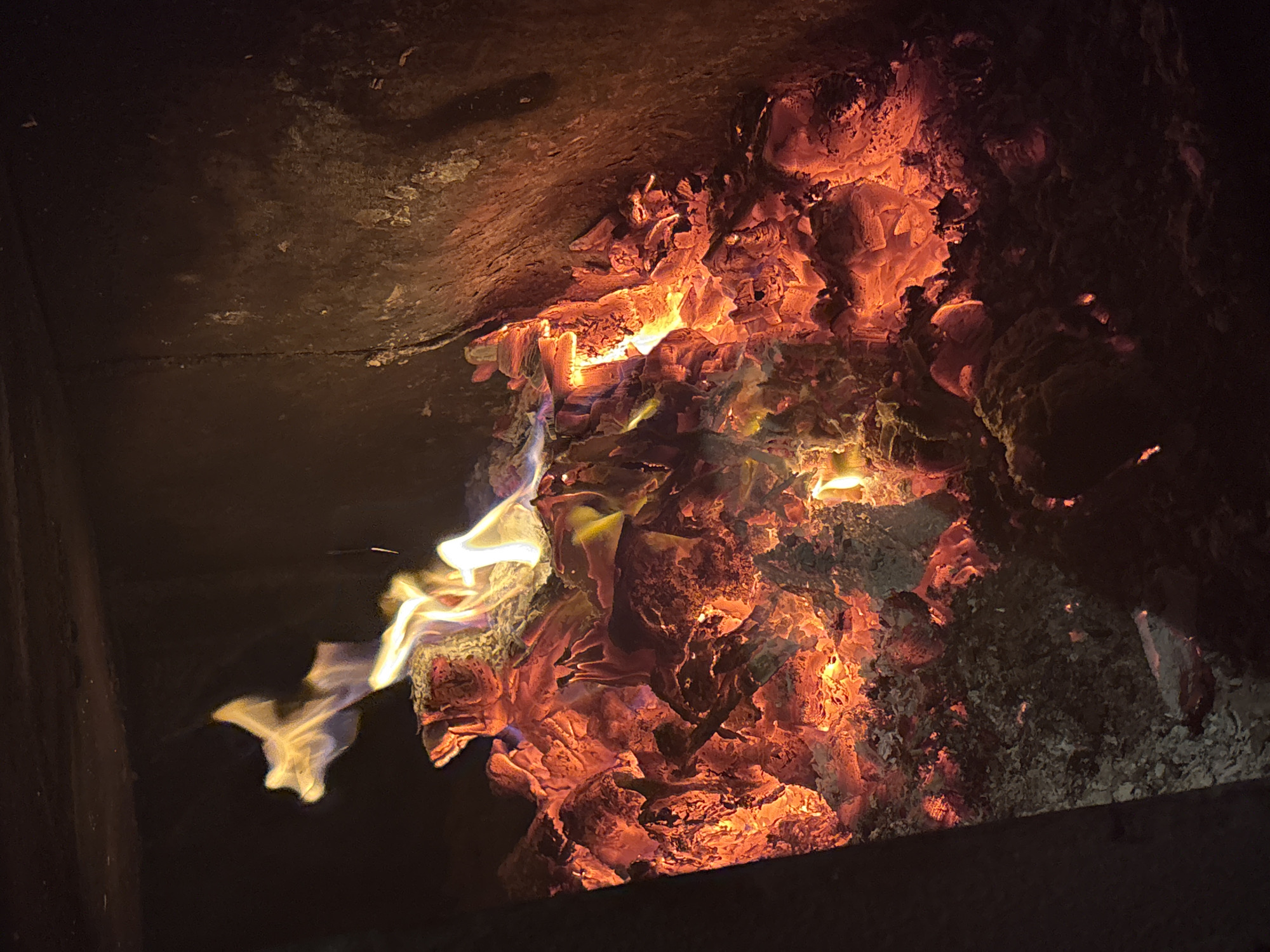 A close-up, vertical shot captures the intense heat and vibrant colors of an active fire burning within a dark, possibly a fireplace or wood-burning stove. The main subjects are the glowing embers and flames. The embers are predominantly a bright, fiery red and orange, with some areas showing lighter yellow and white hues where the heat is most concentrated. These embers form a dense, uneven pile, suggesting partially burned logs and ash.
Flames, depicted in swirling shapes of bright yellow and white, rise from the embers, illuminating the scene. Some wisps of flame are tinged with blue and purple, hinting at the chemical reactions occurring during combustion. The textures of the embers range from coarse and granular ash to smoother, more molten-looking surfaces.
The surrounding environment is dark and appears to be the interior of a firebox. To the right, a dark, rough texture, likely a stone or metal wall, is visible, blackened and possibly stained by soot. To the left, a dark, solid structure, possibly another wall or the edge of the firebox opening, is also present. The overall lighting is dominated by the fire itself, creating a stark contrast between the bright, warm core and the surrounding dark, cool areas. The image conveys a sense of raw energy and heat.
There is no indication of the city of Курган, Russia, or any specific geographic location within the image itself. The scene is entirely focused on the fire. There are no people, animals, or any other identifiable objects besides the fire and its immediate surroundings within the firebox. The action is solely the burning of the fuel.