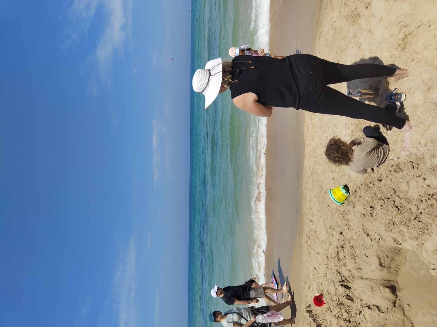 The image depicts a bright, sunny day at a beach. The sky is a clear, vibrant blue with scattered wisps of clouds. The ocean stretches out, displaying shades of turquoise and green near the shore where gentle waves break.
On the sandy beach, a woman in a black outfit and a wide-brimmed white hat is positioned near the water's edge. A child, wearing a tan-colored shirt, sits near her, seemingly engrossed in playing in the sand. A yellow and green toy bucket is near the child.
Further down the beach, closer to the bottom of the frame, are three adults and one child, possibly interacting. A small red bucket lies nearby.
The scene appears to be at a beach in Tel-Aviv, Israel, evidenced by the ocean, the beach, and the overall atmosphere.