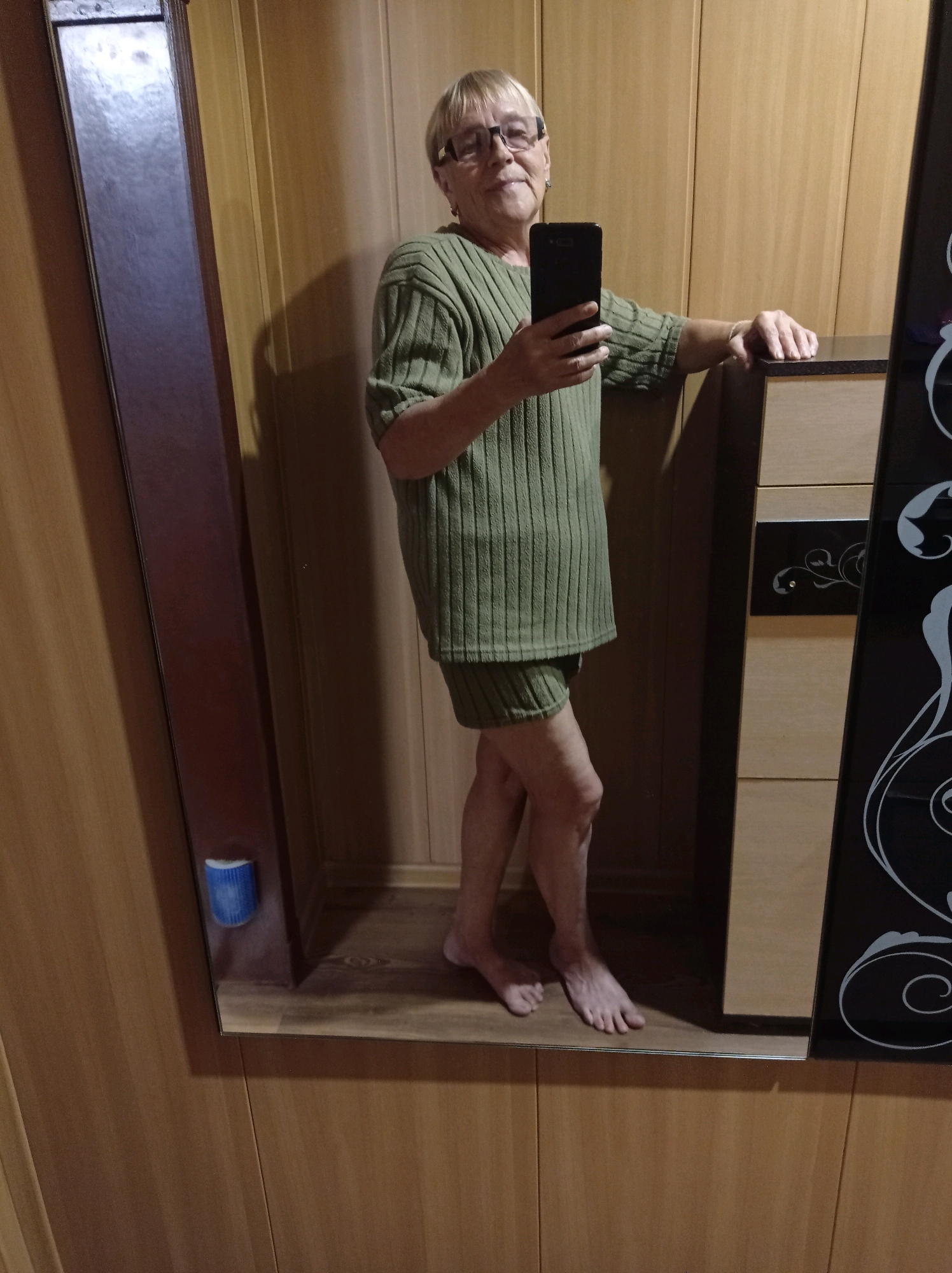 A full-body mirror selfie shows a person standing in what appears to be a bedroom or dressing area. The person is wearing a green ribbed sweater and matching shorts, and is holding a black smartphone in their right hand. They are looking at the camera with a slight smile. The background features wooden walls and a large mirror that reflects the scene. There is also a piece of furniture with drawers to the right of the person. The floor appears to be wooden. The image is taken indoors, and the lighting suggests it is daytime. There is no specific indication of the location within the city of Degtyarsk, Russia, beyond it being an indoor residential setting.