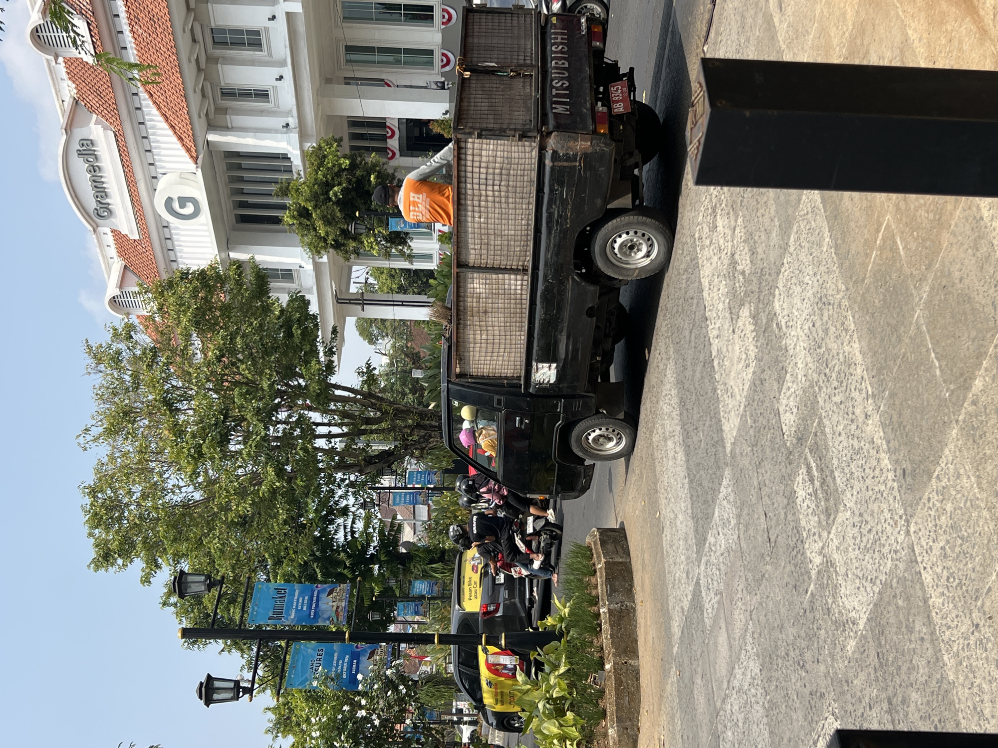 The image captures a street scene in Yogyakarta, Indonesia, with the distinctive white building of Gramedia bookstore featuring a large "G" logo prominently in the background. In the foreground, a black Mitsubishi pickup truck is moving along the street. A person wearing an orange shirt is seated in the back of the truck, facing away from the camera. The truck bed contains a broom and other assorted items. The street is paved with textured gray and white tiles. Several motorcycles are visible on the road, along with a black car branded with yellow text. Banners hang from lampposts, one of which displays the word "Rumaket." The overall environment suggests a bustling urban setting on a clear day.