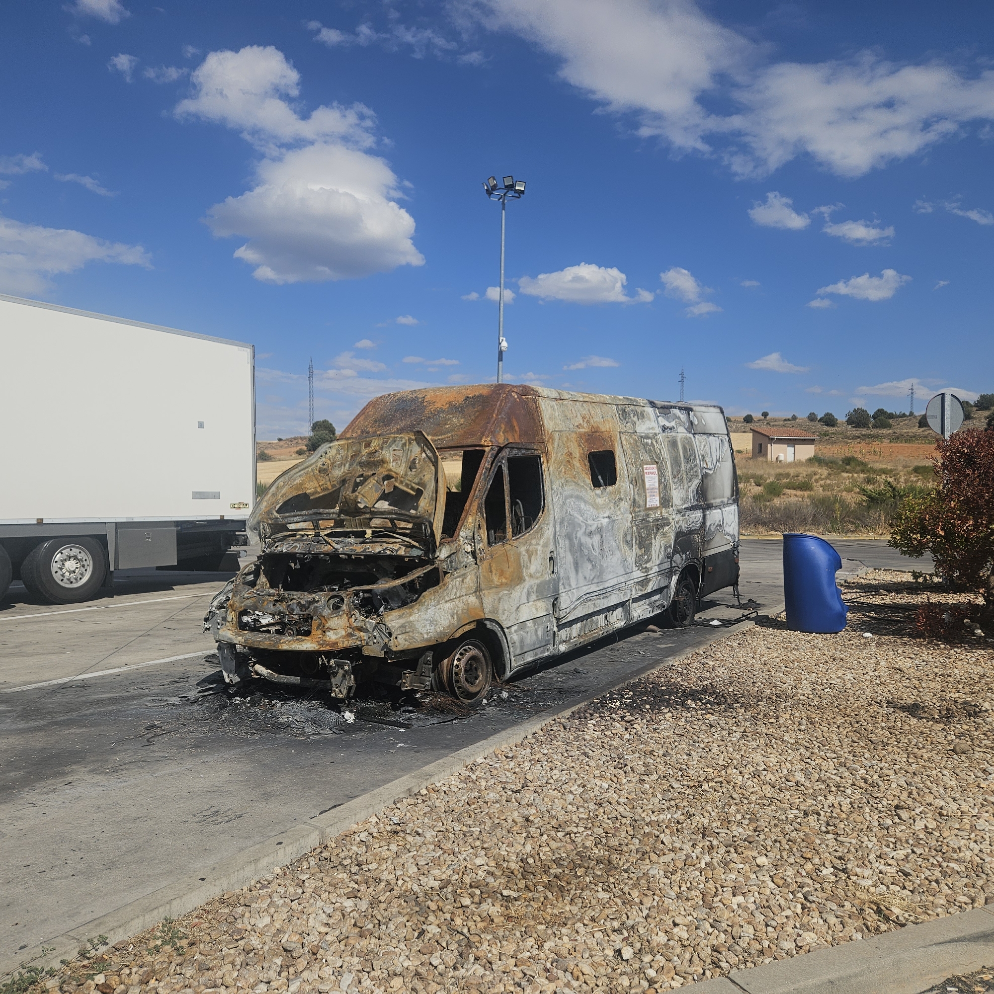 The image depicts a burnt-out van situated on the shoulder of a road, adjacent to a white trailer truck. The van's exterior is charred and melted, with its front end heavily damaged and its tires missing or melted. The interior of the van is visible through the broken windows and gaping holes. A blue cylindrical object, possibly a trash receptacle, is located to the right of the van. The scene is set against a backdrop of a bright blue sky with scattered white clouds, dry vegetation, and a small building in the distance. The location is identified as being near El Burgo de Osma-Ciudad de Osma, Spain, suggested by the presence of similar landscapes and infrastructure in other images from that region. There are no people visible in the image.