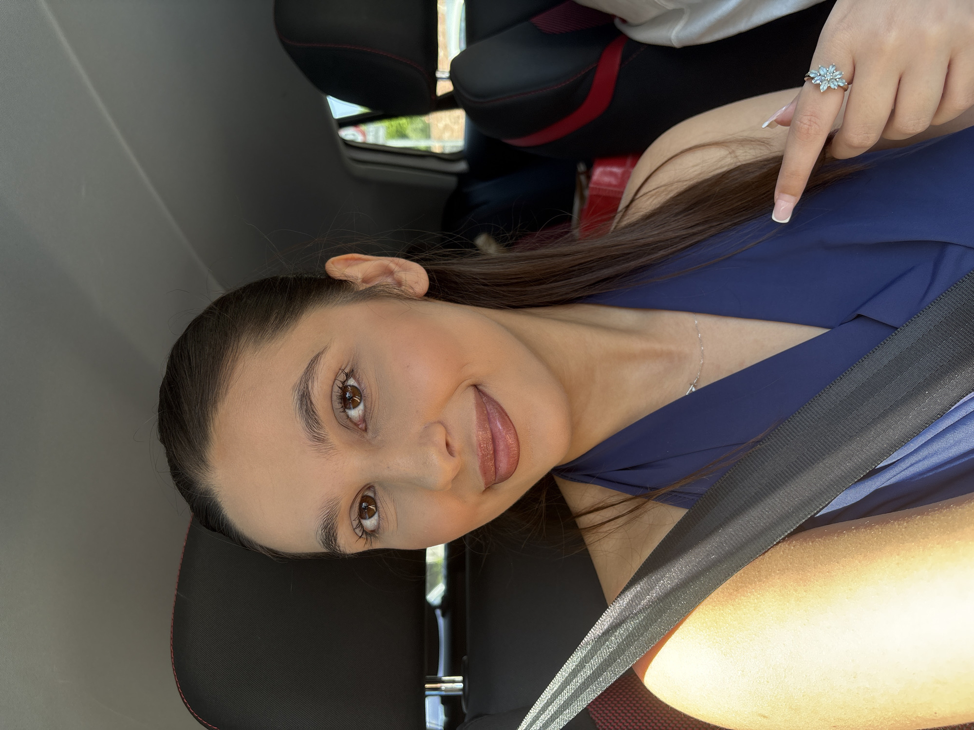 A woman with long brown hair, wearing a blue top and a seatbelt, is seated in a vehicle, likely a car, with black seats with red accents. She is looking at the camera with a slight smile. Another person's hand with a floral-shaped ring is also visible in the upper right. The setting is a car interior; the location is not discernible within Mönchengladbach, Germany, from this image.
