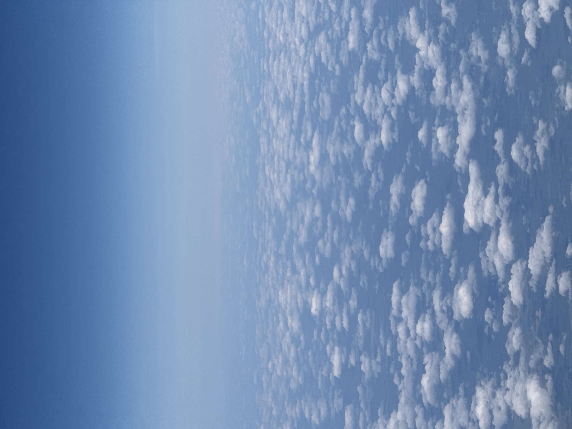 The image depicts a vast expanse of clouds seen from above, likely from an airplane window. The sky is a gradient of blues, transitioning from a deep, clear blue at the top to a lighter, hazy blue towards the horizon. Below the sky, a dense layer of white, puffy cumulus clouds stretches across the frame, creating a textured, undulating surface. The clouds appear to be at a considerable altitude, obscuring any view of the ground beneath. There are no people, specific objects, or actions visible in the image, only the natural phenomenon of clouds in the atmosphere. The location in the city of Kazan, Russia, is not discernible from the visual information provided in the image.
