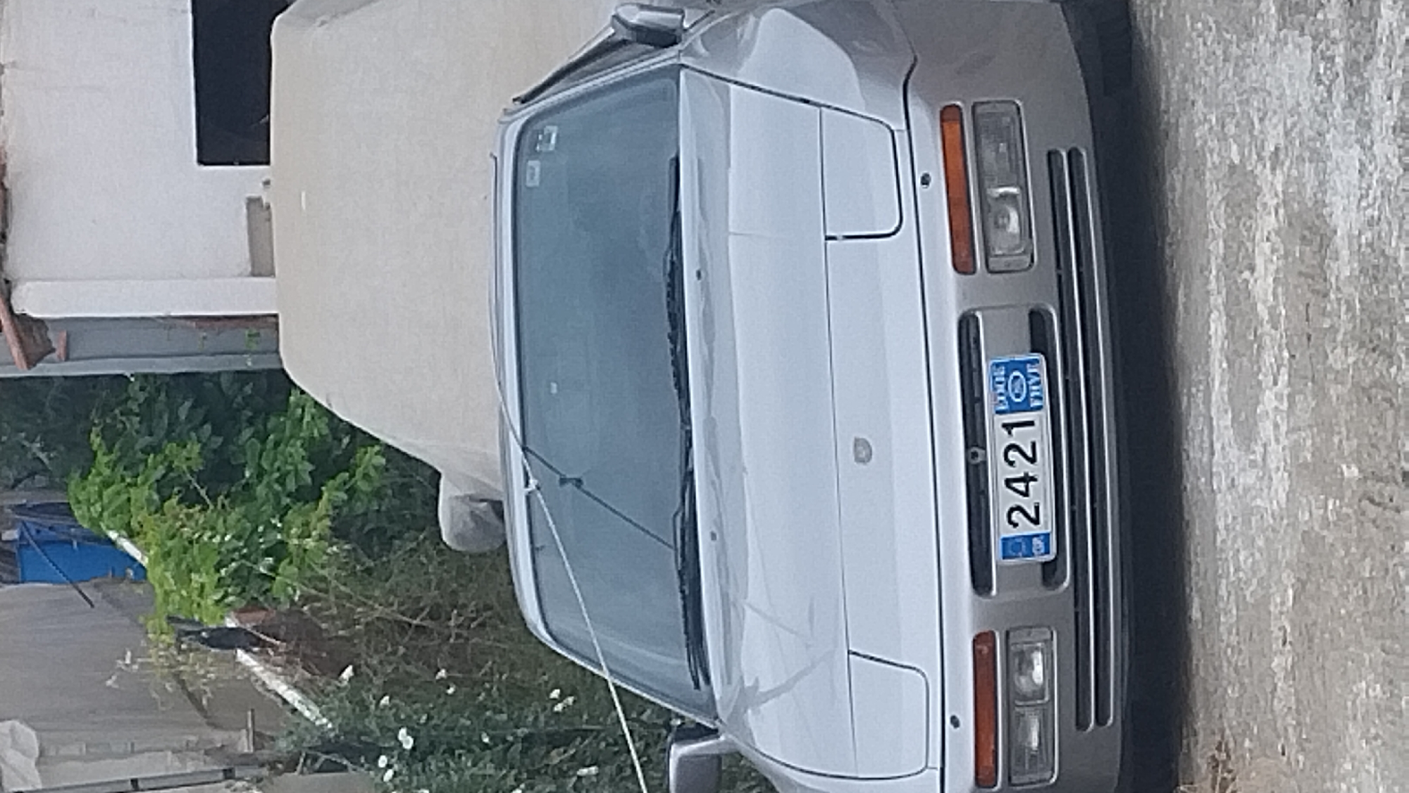 The image displays the front of a silver Porsche 944, parked outdoors in an urban or semi-urban setting, possibly in Νέα Κερασιά, Greece, given the context. The car's pop-up headlights are down. A blue and white license plate is visible on the front bumper, reading "2421". The car is partially covered by a light-colored tarp. In the background, there are dense green bushes and a white building with a dark opening, suggesting a residential or storage area. The ground in front of the car is paved with rough, grey material.