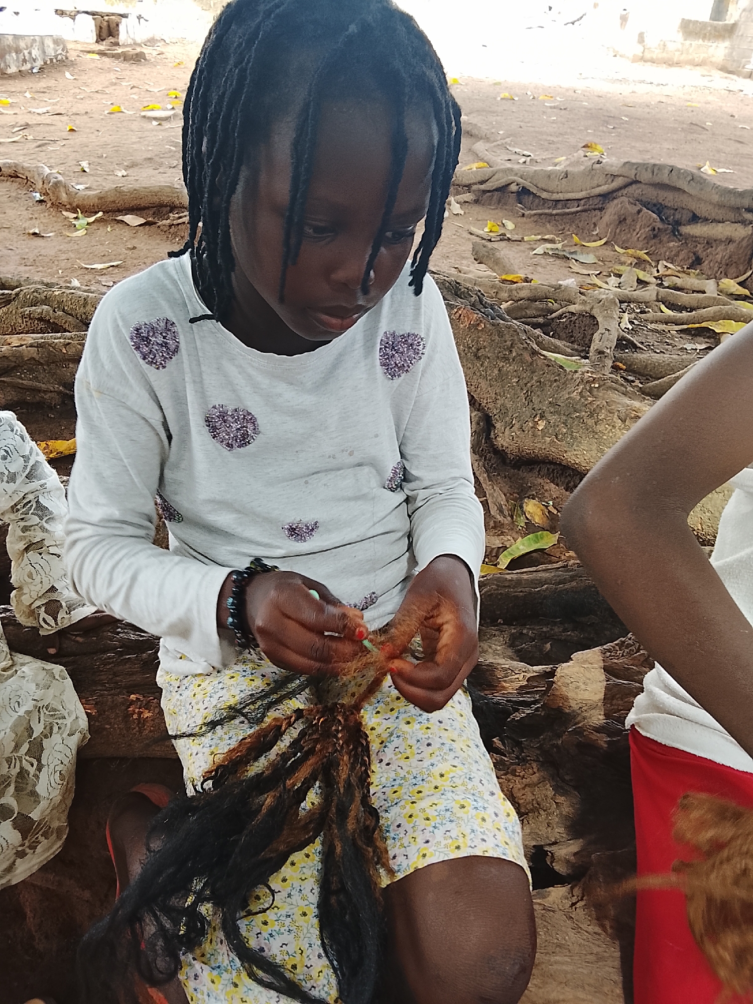A young Black girl is sitting outside on what appears to be tree roots or a similar natural surface. She is wearing a long-sleeved light grey shirt with purple patterned circles, and a yellow floral skirt. She is holding and working with brown and black hair extensions or dreadlocks. The location is in the city of Unknown, Unknown, though no specific landmarks can be seen. Another person's arm can be seen to the right, possibly assisting her. The background consists of an outdoor setting, possibly a yard or park.